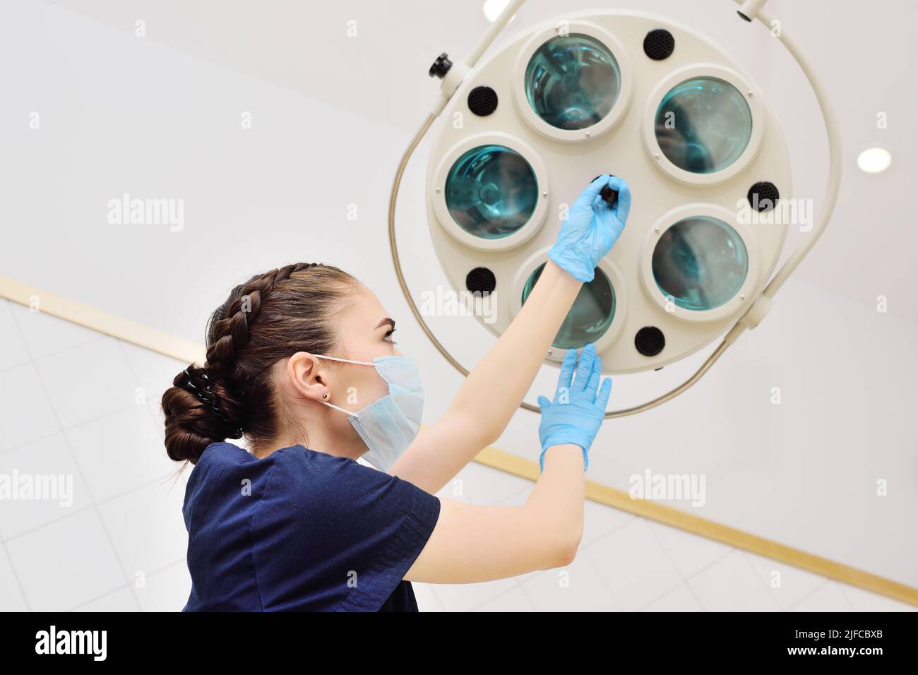 Attraktive weibliche Sanitäter in einer sterilen Maske und Gummihandschuhen schaltet eine Operationslampe in einem modernen Operationssaal ein. Stockfoto