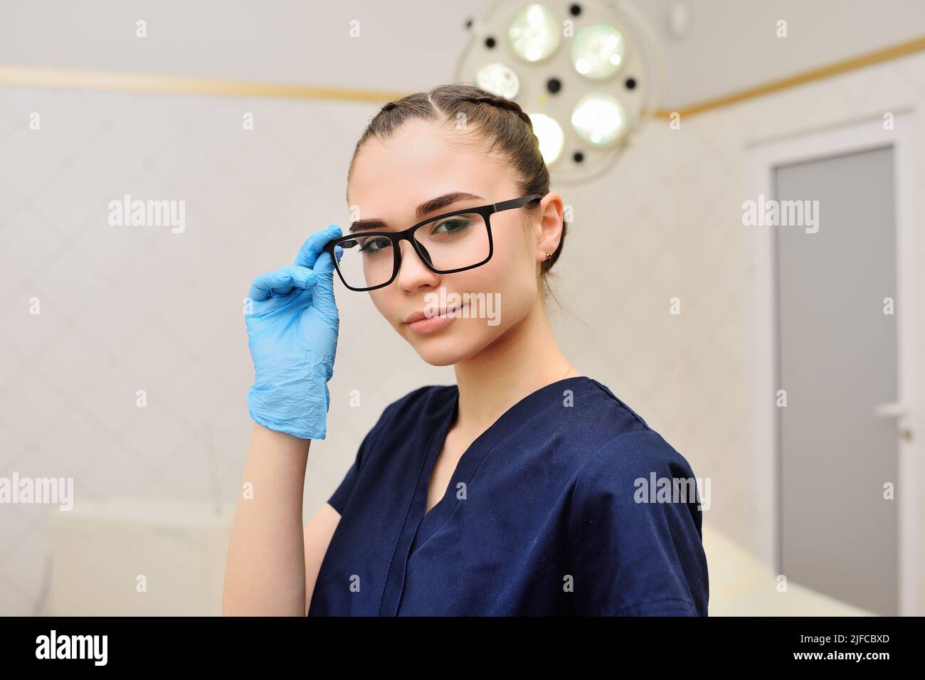 Junge Ärztin in Gummihandschuhen lächelt und stellt die Brille vor den Hintergrund einer Operationslampe Stockfoto