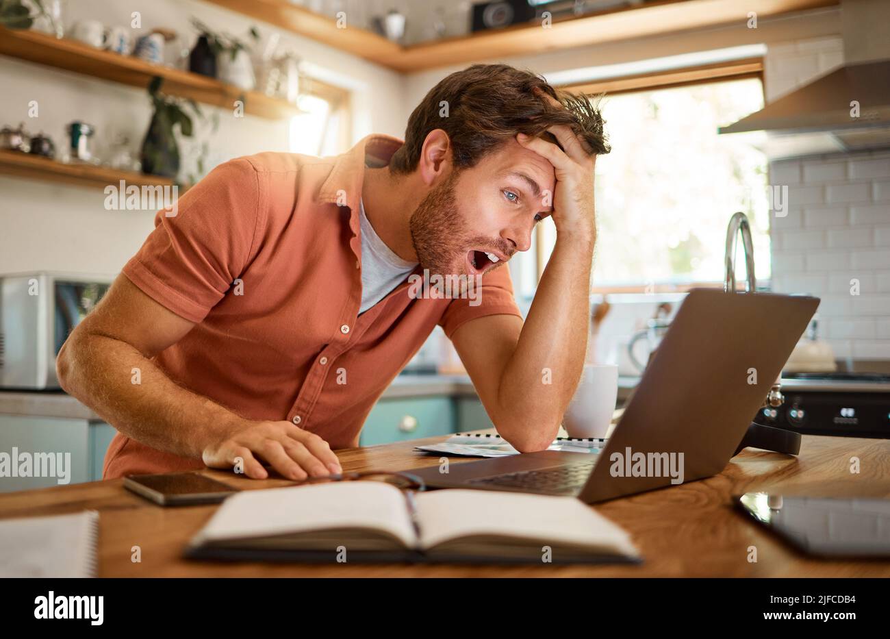 Junger, verblüffender kaukasischer Geschäftsmann, der allein zu Hause an einem Laptop arbeitet. Ein schockierter männlicher Geschäftsmann sah überrascht aus, als er in der Küche arbeitete Stockfoto