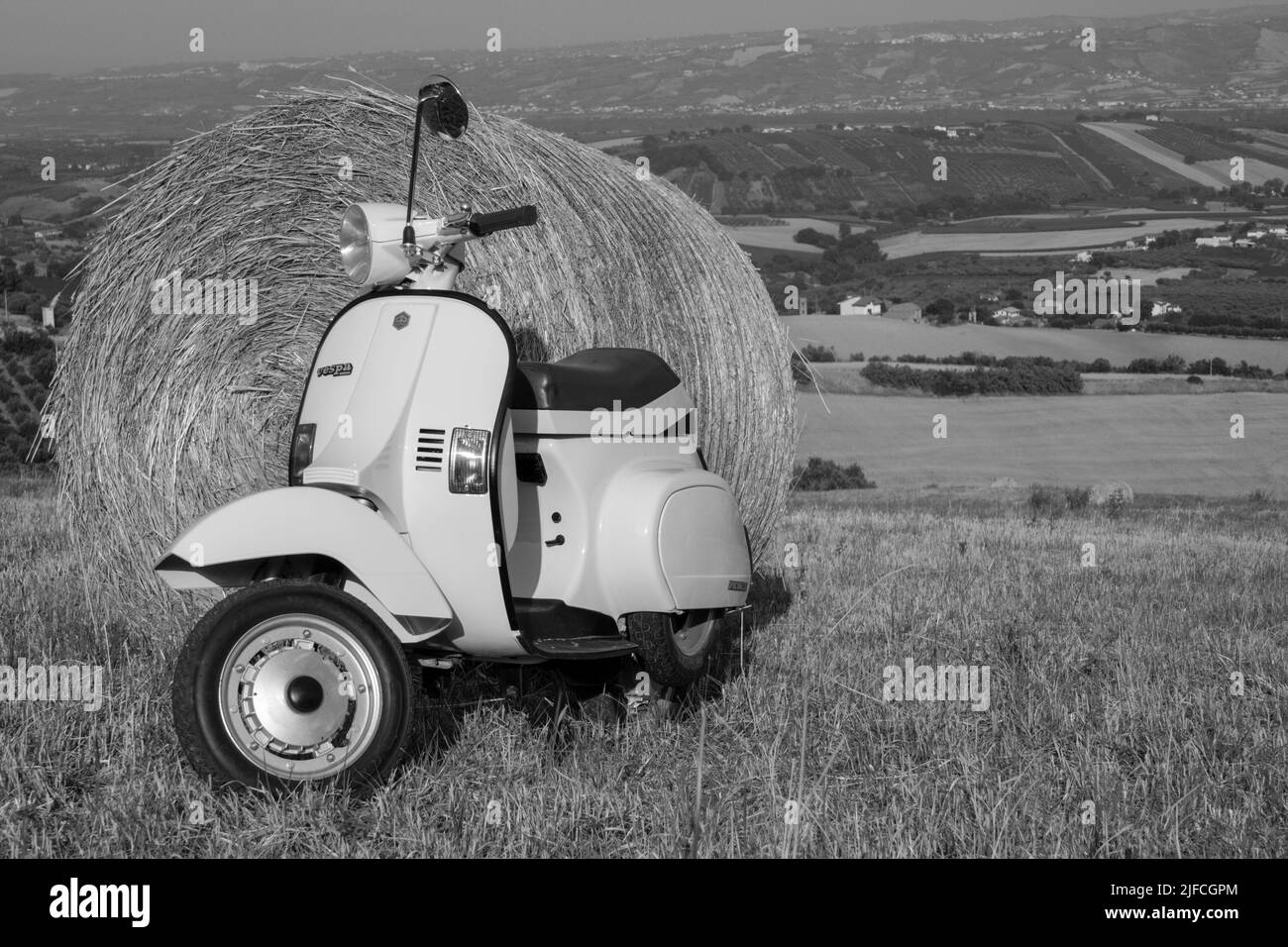 Bild eines alten Piaggio vespa Motorrads, das auf einem Feld mit Strohballen und einer atemberaubenden Aussicht im Hintergrund geparkt ist. Schwarzweißfotos. Stockfoto