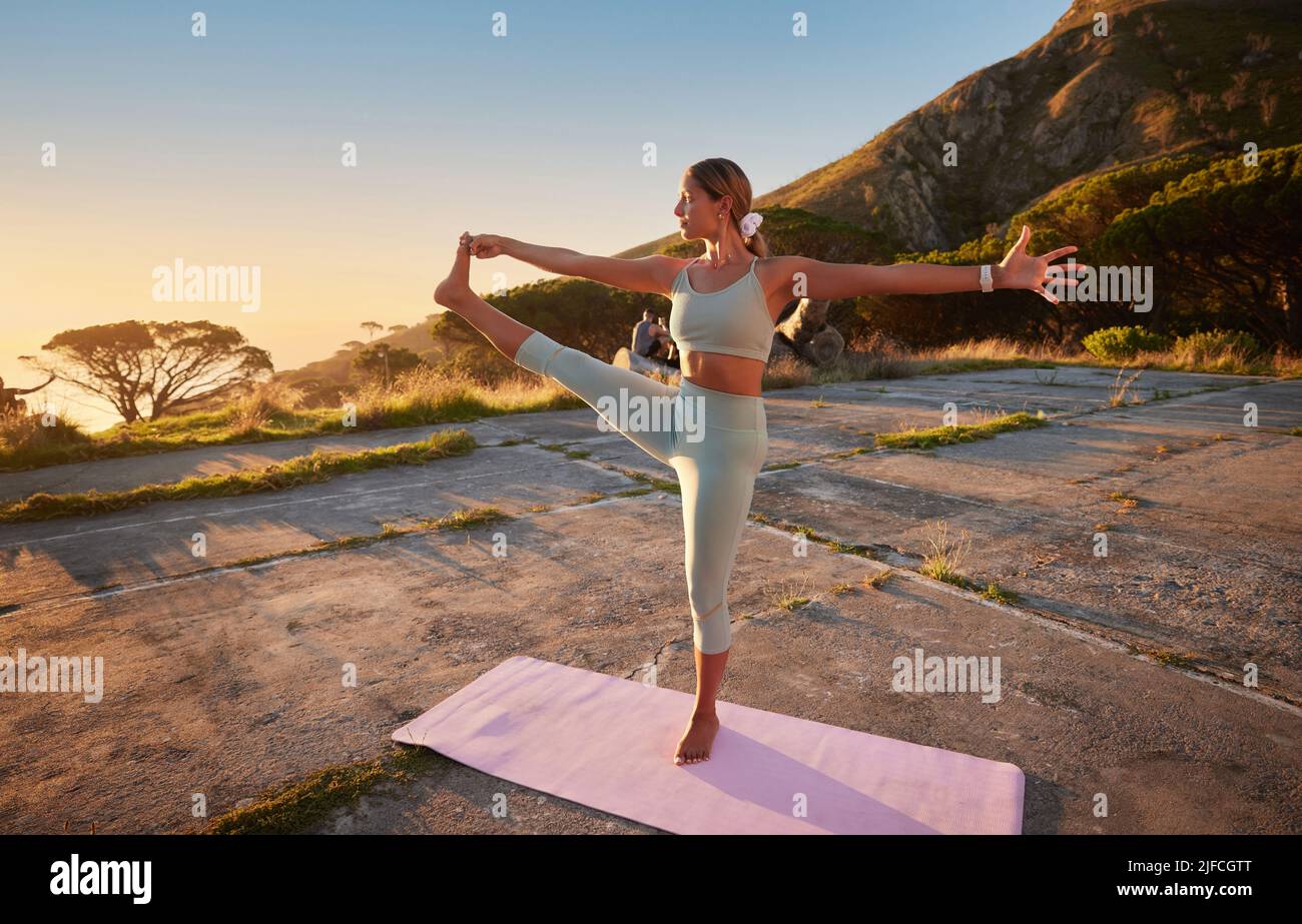 Yoga-Frau in voller Länge, die Hand an Fuß hält, in der Natur im Freien üben. Schöne kaukasische Person mit Matte, balancieren beim Dehnen Stockfoto