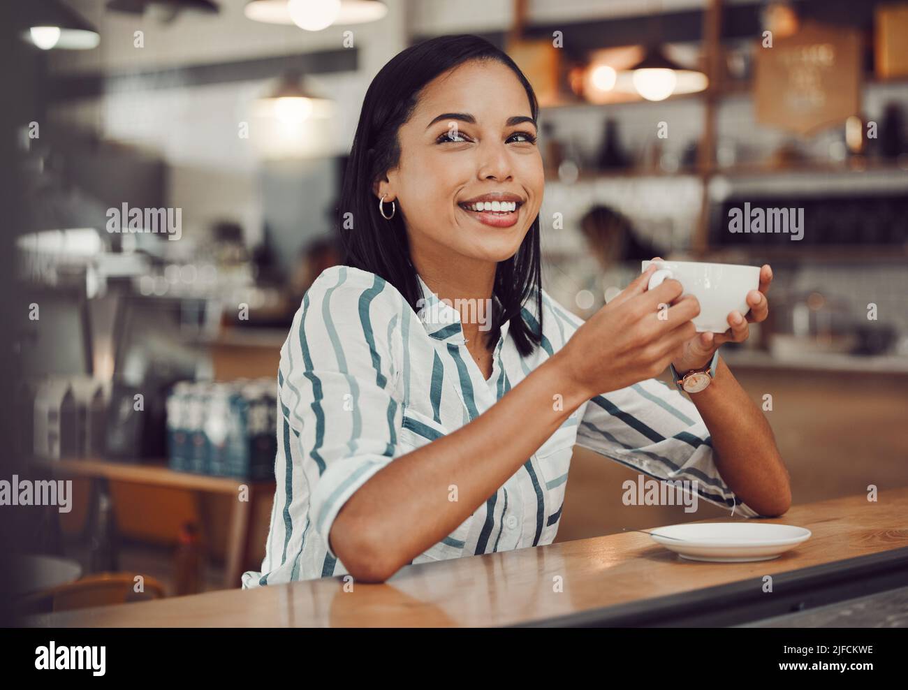 Fröhliche junge lächelnde Mischling Frau im lässigen Outfit, die heißes Getränk trinkt, während sie allein an einem Tisch im gemütlichen Café sitzt. Schöne weibliche aussehende Stockfoto