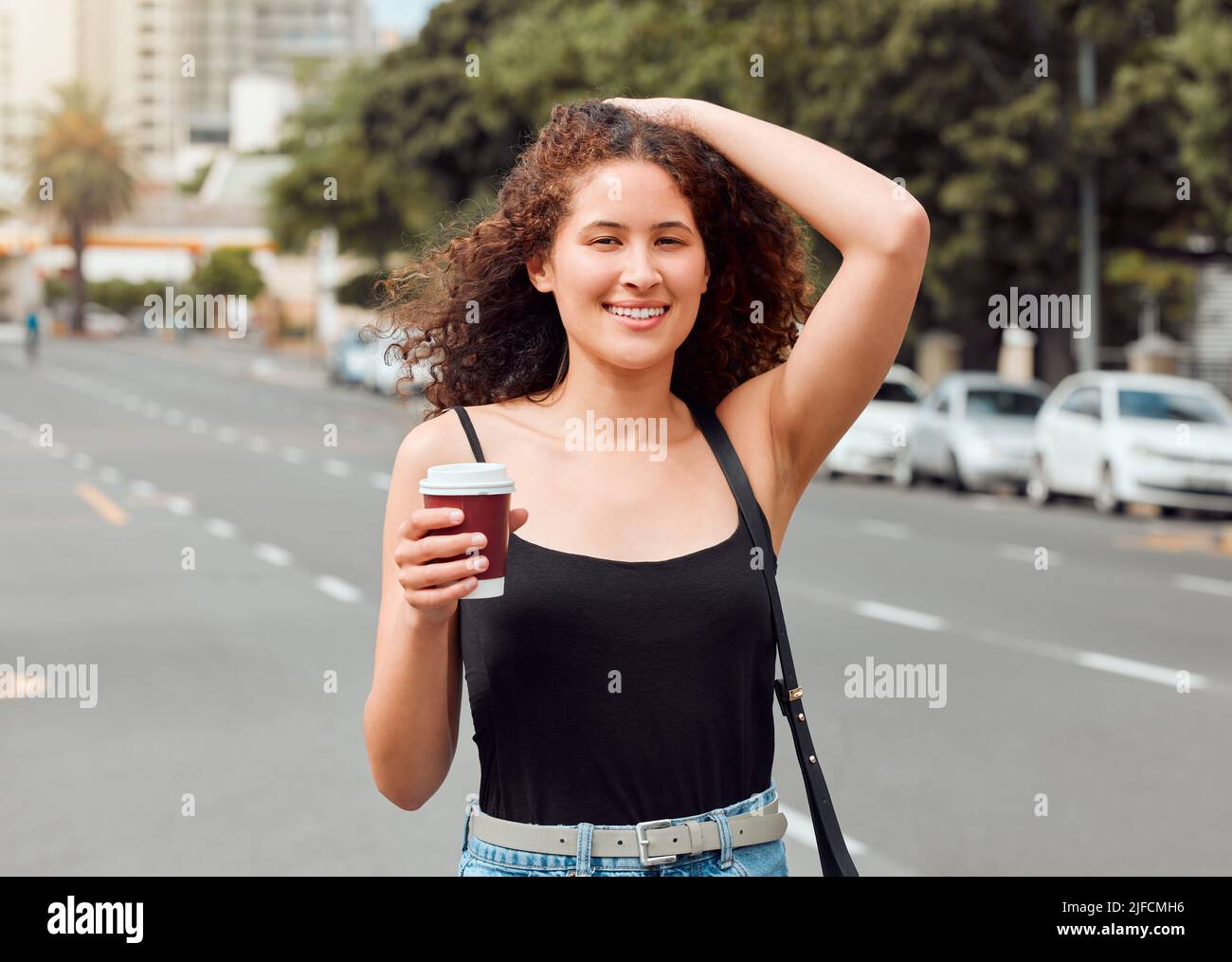 Porträt einer glücklichen, schönen jungen Frau mit gemischter Rasse, die die Stadt erkundet, während sie einen Kaffee zum Mitnehmen hält und ihr lockiges, brüniertes Haar berührt. Hispanisch Stockfoto