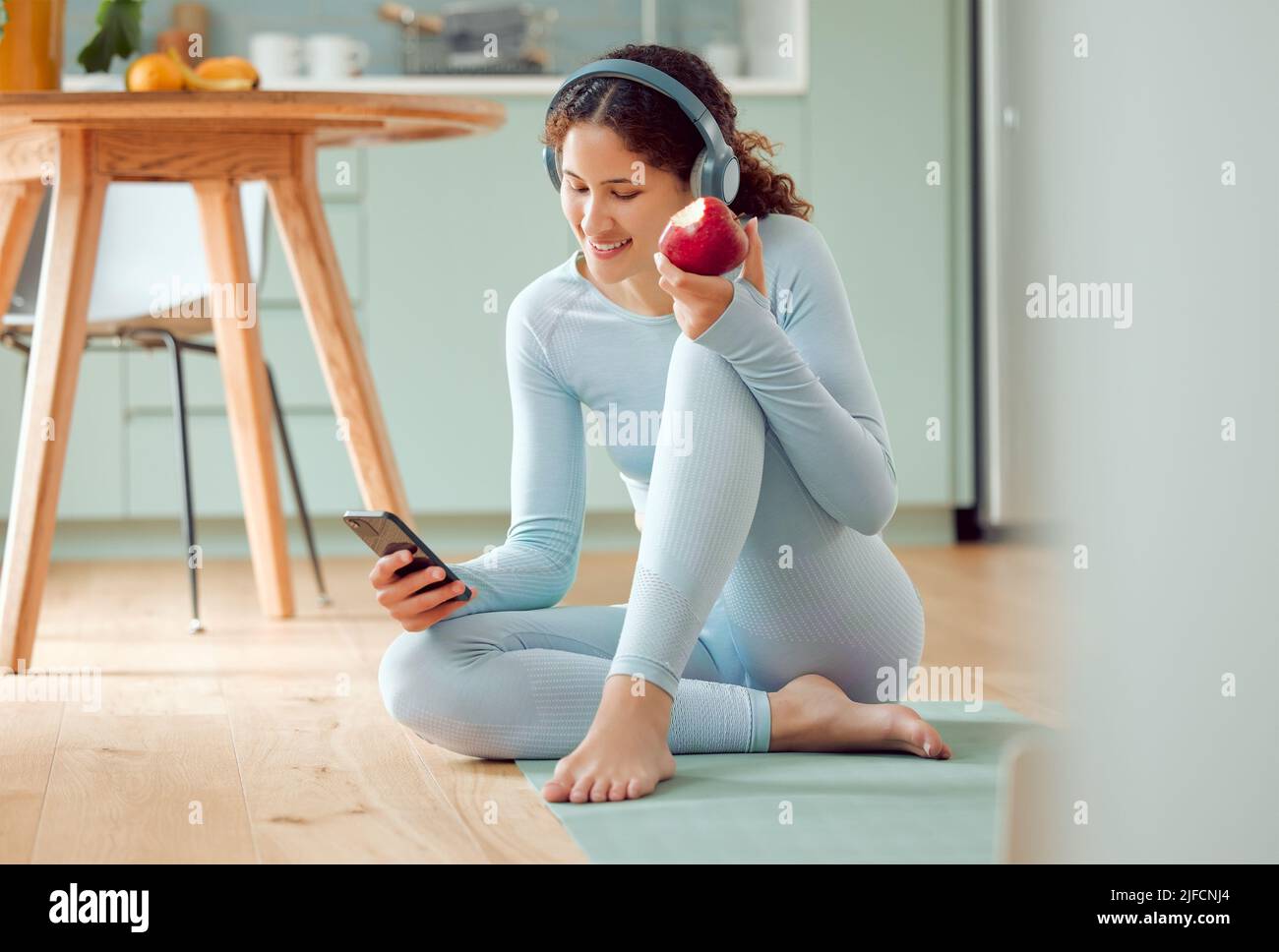 Schöne junge Frau mit gemischter Rasse, die Musik hört, während sie zu Hause eine Pause vom Yoga macht. Hispanische Frau, die ihr Telefon benutzt und eine isst Stockfoto
