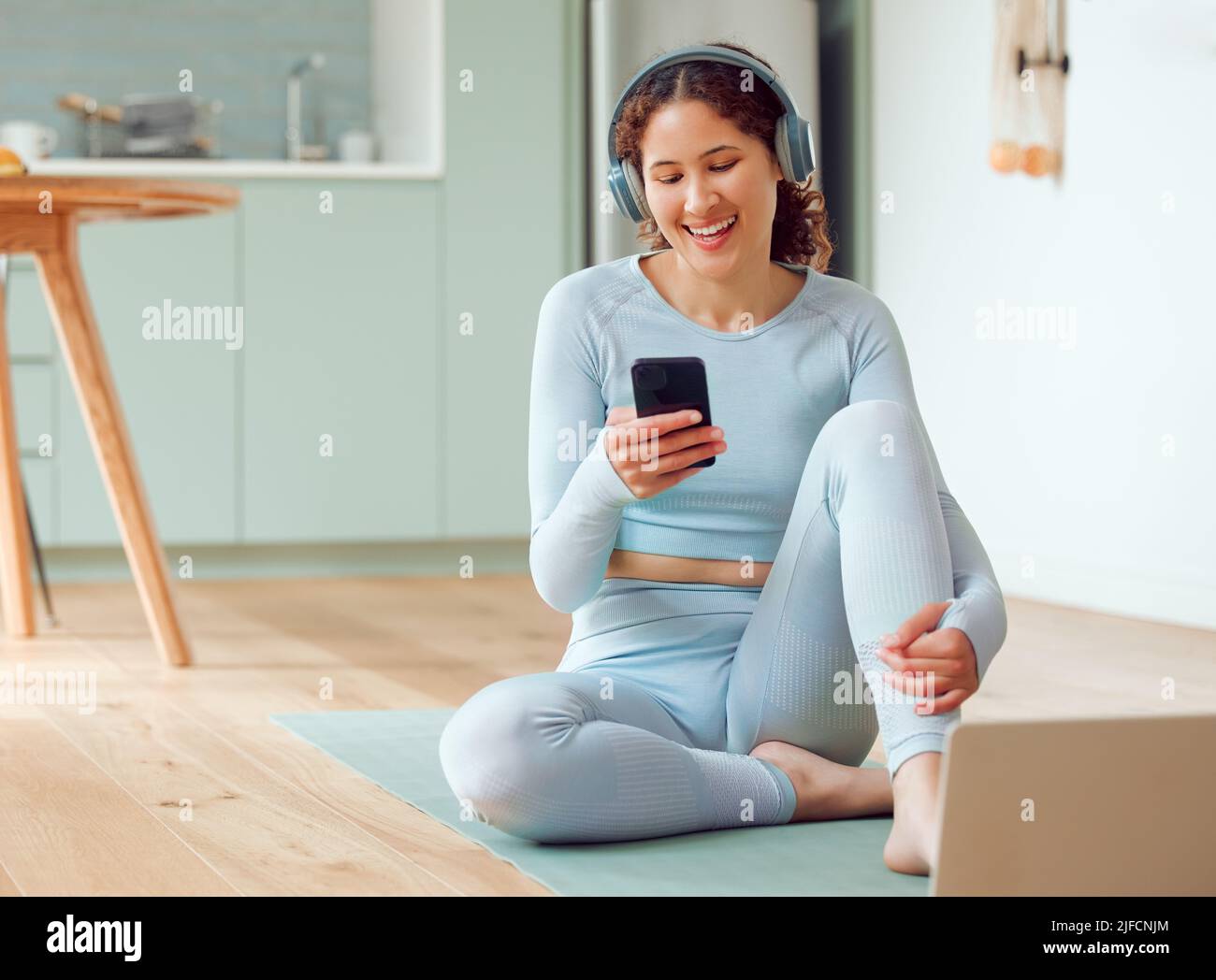 Schöne junge Frau mit gemischter Rasse, die Musik hört, während sie zu Hause eine Pause vom Yoga macht. Hispanische Frau, die ihr Telefon benutzt, um eine SMS zu senden Stockfoto