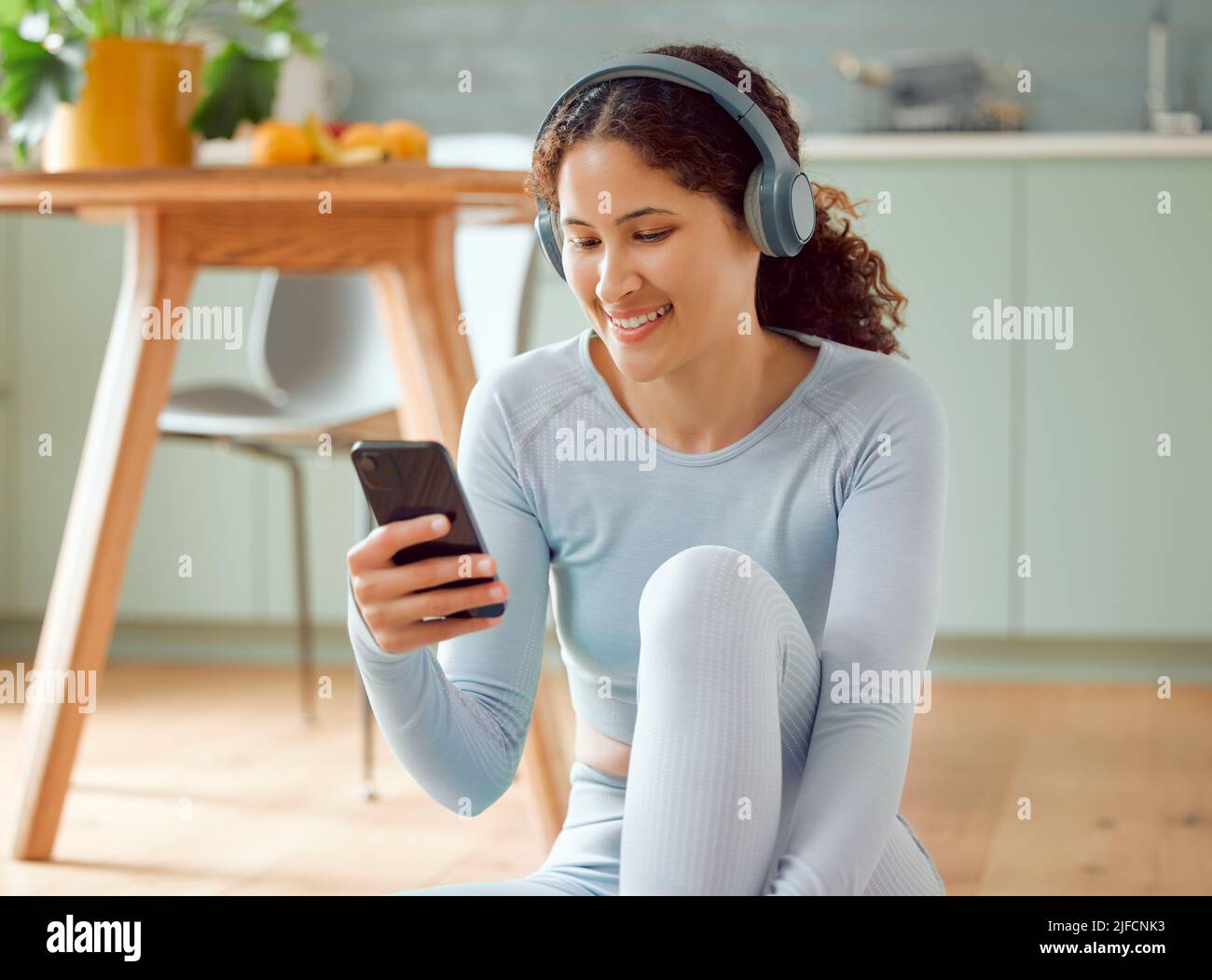 Schöne junge Frau mit gemischter Rasse, die Musik hört, während sie zu Hause eine Pause vom Yoga macht. Hispanische Frau, die ihr Telefon benutzt, um eine SMS zu senden Stockfoto