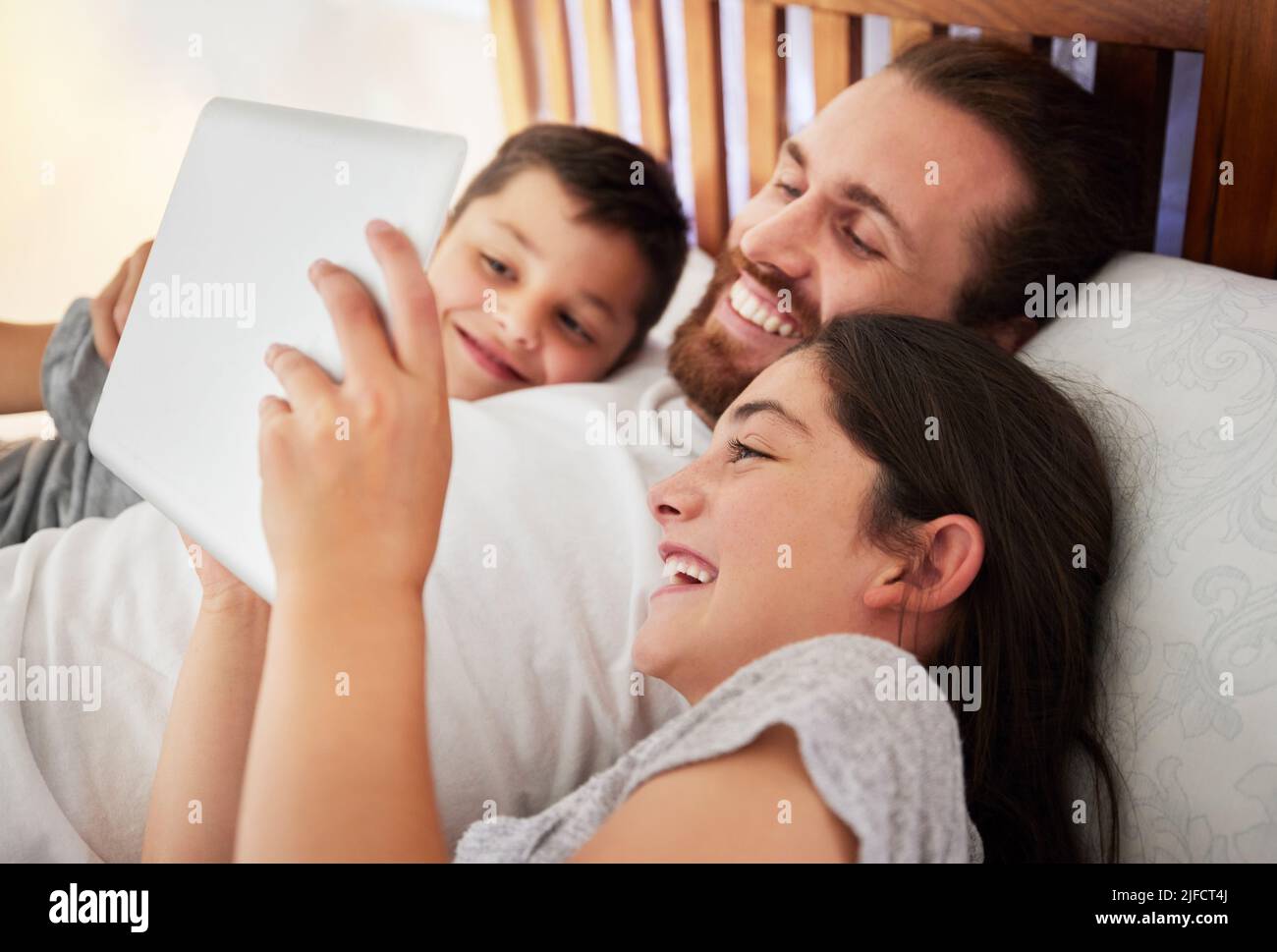 Glücklicher kaukasischer Vater, der mit seinen beiden Kindern im Bett liegt und Filme ansieht, Online-Bücher liest oder zusammen Spiele spielt Stockfoto