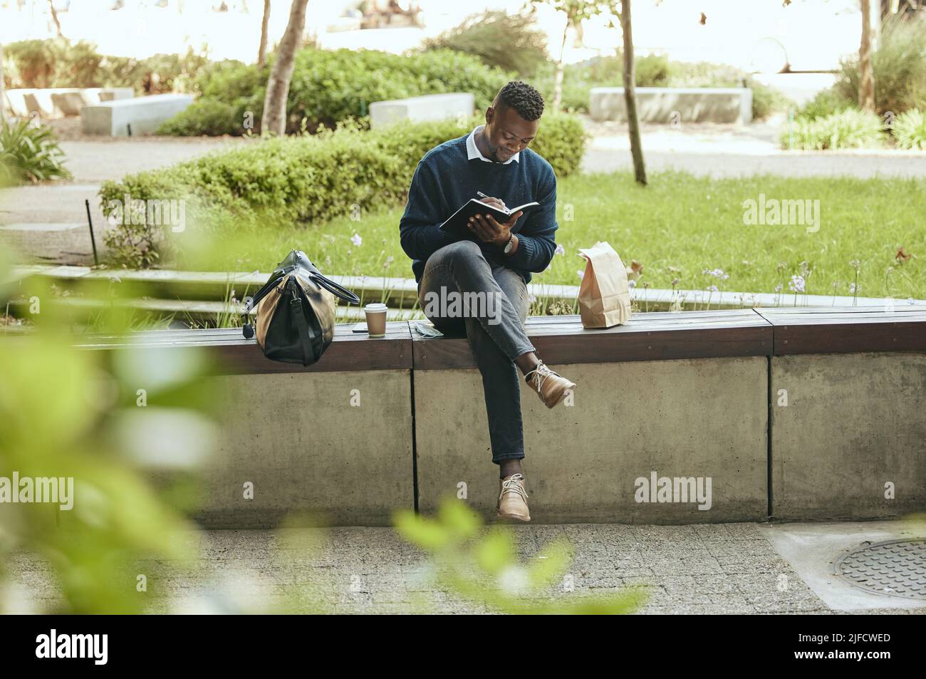 Schwarzer Geschäftsmann, der in einem Notizbuch schreibt, während er in einem Park sitzt. Eine Mittagspause vom Büro, um etwas Planung in seinem Tagebuch zu machen Stockfoto