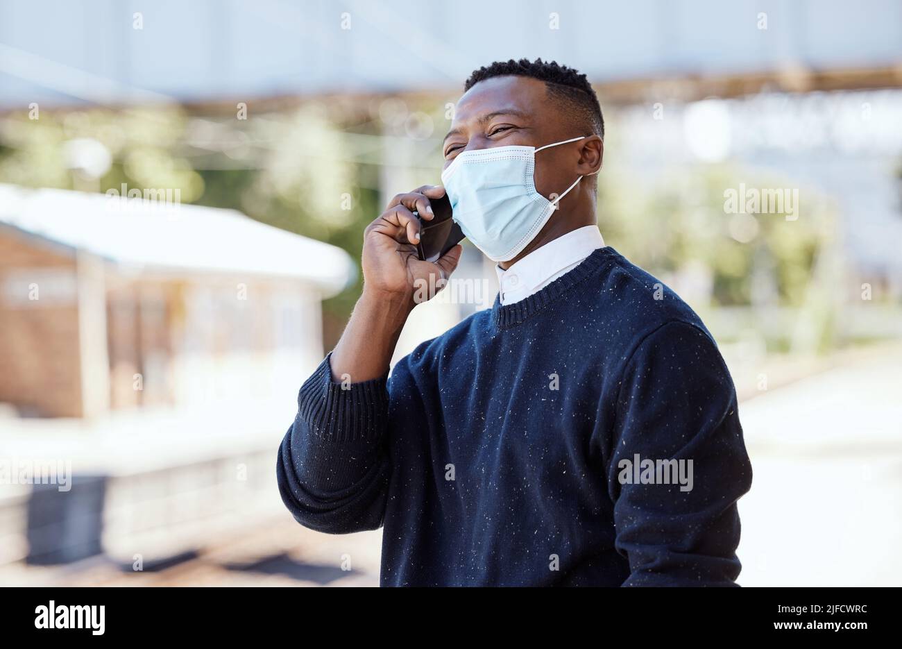 Schwarzer Geschäftsmann, der eine Gesichtsmaske trägt und draußen auf einem Handy spricht. Während der covid pandemic einen Anruf tätigen, während der Pendelverkehr ansteht Stockfoto