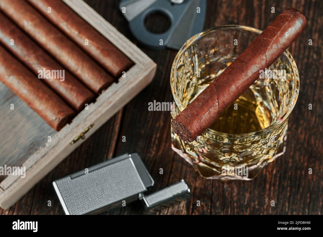 Blick von oben auf eine kubanische Zigarre auf einem Glas Whiskey oder Alkohol. Holztisch, Feuerzeug und Cutter mit unscharfem Hintergrund. Stockfoto