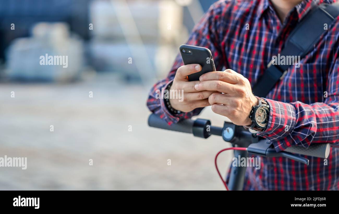 Junger Mann mit Hemd, der sich am Lenker des Elektrorollers lehnt, hält das Smartphone in den Händen, Nahaufnahme, Platz für Text links Stockfoto