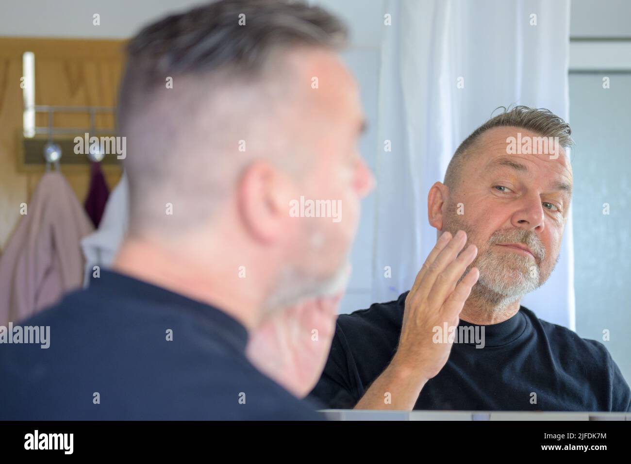 Ein älterer Mann, der seine Gesichtshaut in einem Spiegel im Badezimmer in einem Anti-Aging- und Hautpflegekonzept überprüft Stockfoto