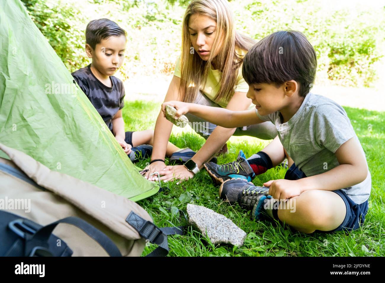 Multiethnische lateinische und kaukasische Kinder nageln Zeltpfeihen auf einem Sommerlager mit einer Lehrerin. Lernen, ein Zelt zu schlagen. Urlaubskonzept Stockfoto