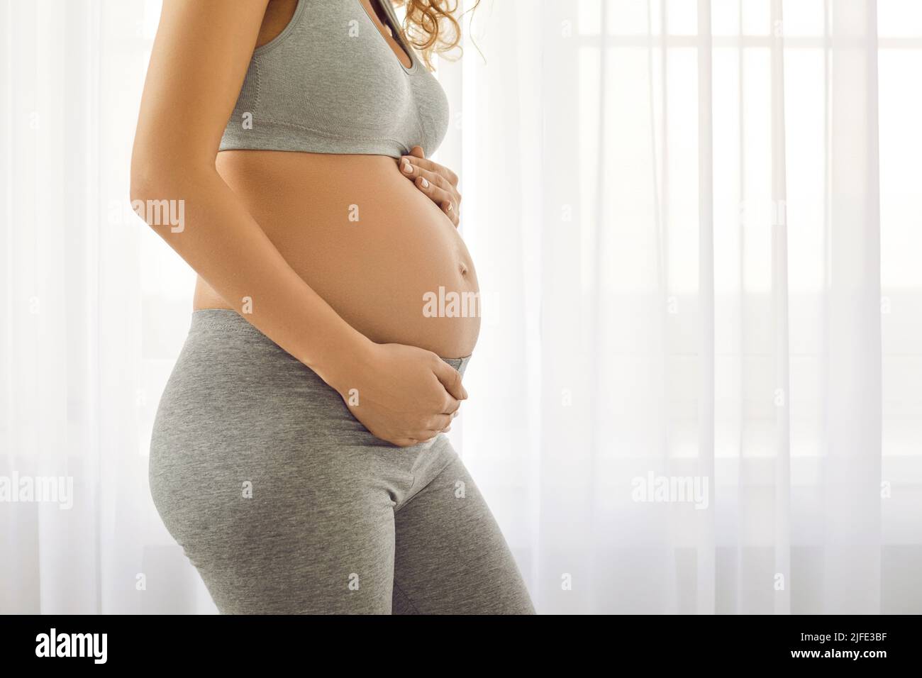 Glückliche werdende Mutter, die vor dem Fensterhintergrund steht und ihren großen Bauch berührt Stockfoto