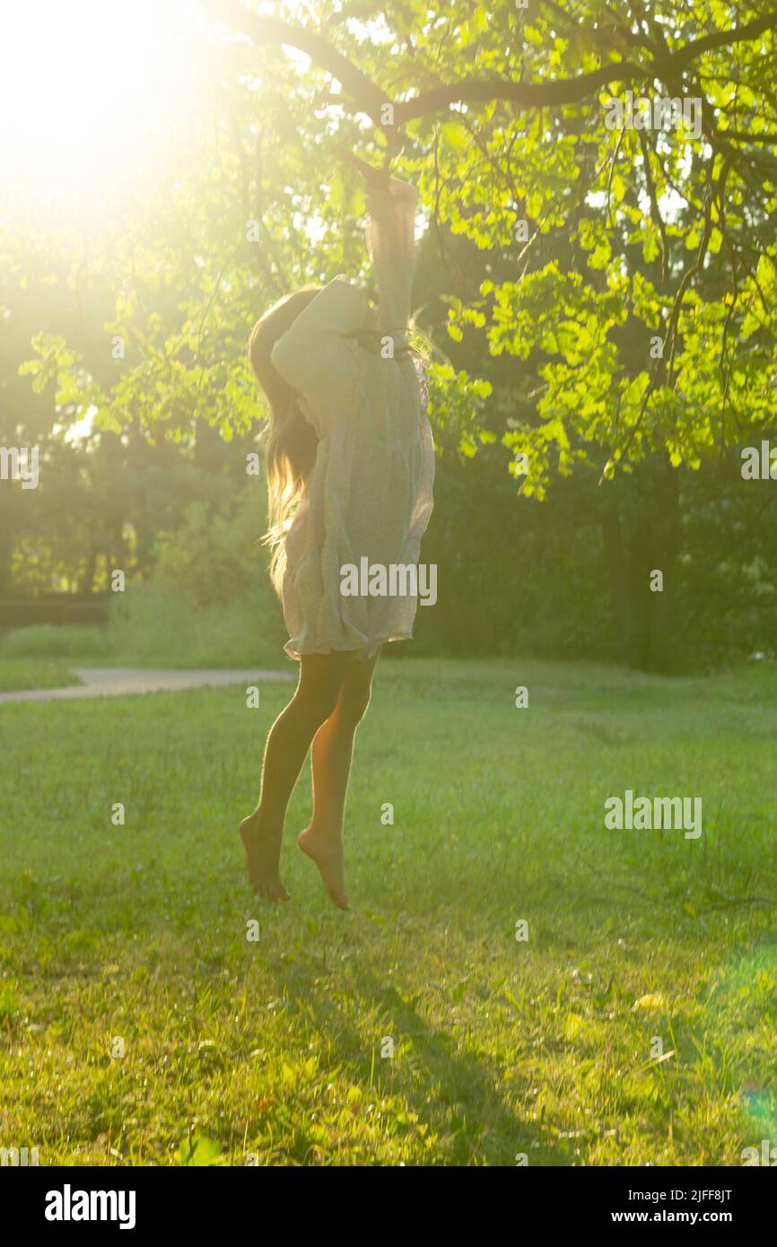 Ein schönes Mädchen in einem Kleid erstreckt sich zu einem Baumzweig im Park. Potenzielles Wachstums- und Zielerreichungskonzept. Stockfoto