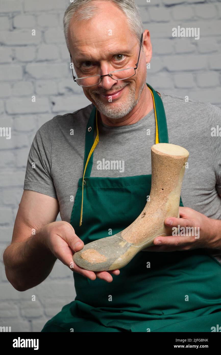 Ein älterer Schuhmacher mit grüner Schürze präsentiert eine individuelle Holzschürze, um einen individuellen orthopädischen Schuh zu fertigen Stockfoto
