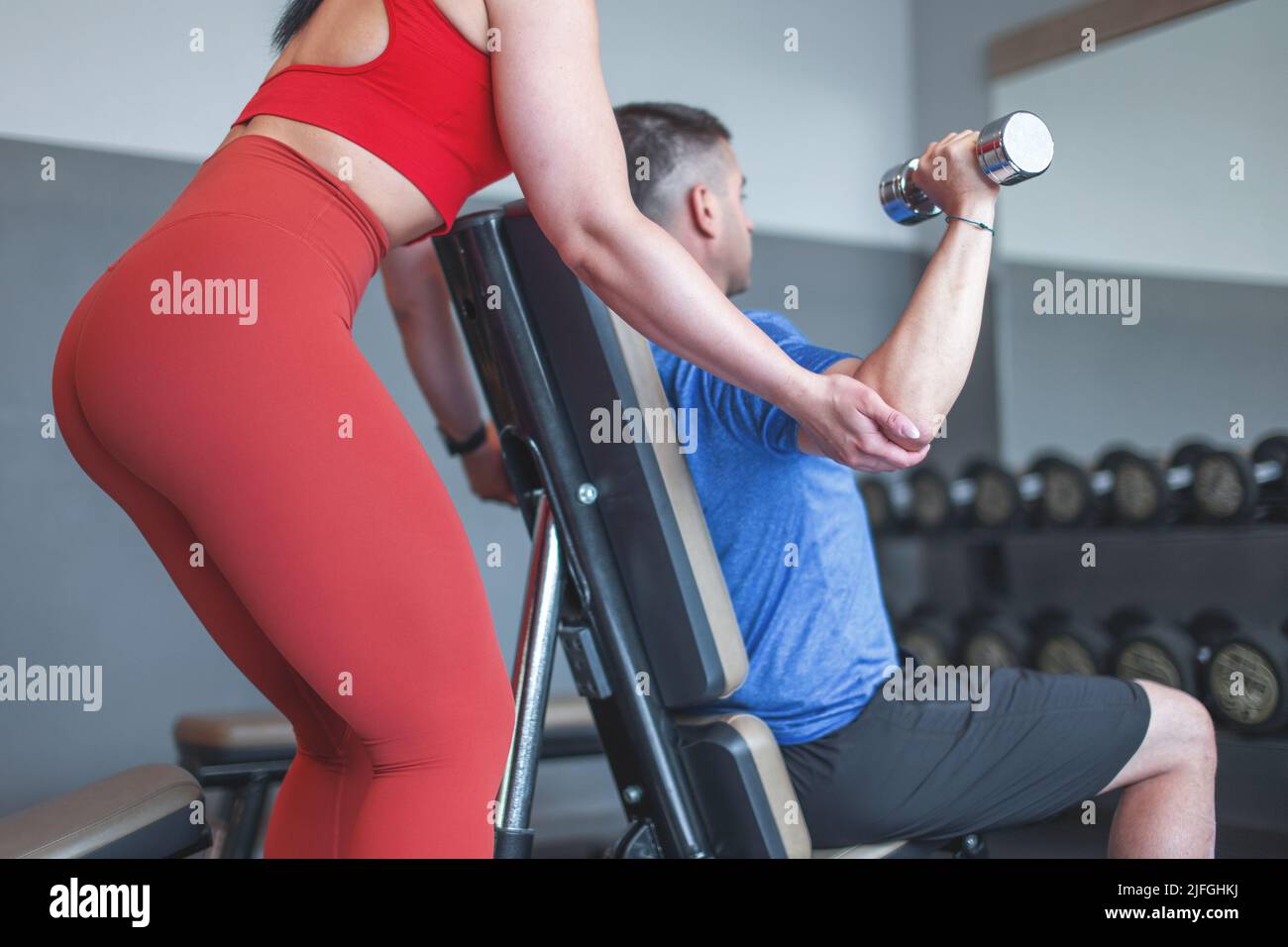 Trainer hilft, das Training des Kunden durch Hanteln im Fitnessstudio am Spiegel zu schwächen Stockfoto