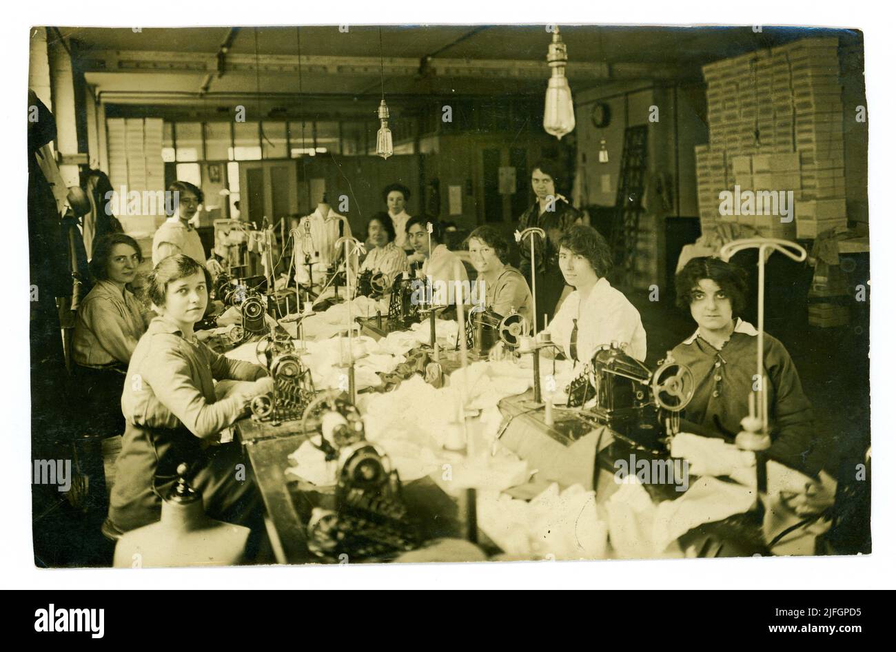 Originalfoto aus der Zeit von WW1 von einer Gruppe junger Arbeiterinnen, die in einer Fabrik arbeiteten, die Blusen mit Tretnähmaschinen herstellte, eine Bluse wird auf einer Schneiderpuppe ausgestellt, jede Dame hat einen Schneideraufsteller, um die fertige Bluse anzuziehen. Eine Managerin schaut in den Hintergrund. Frühe Glühlampen hängen über der Werkbank. Ca. 1916. GROSSBRITANNIEN Stockfoto