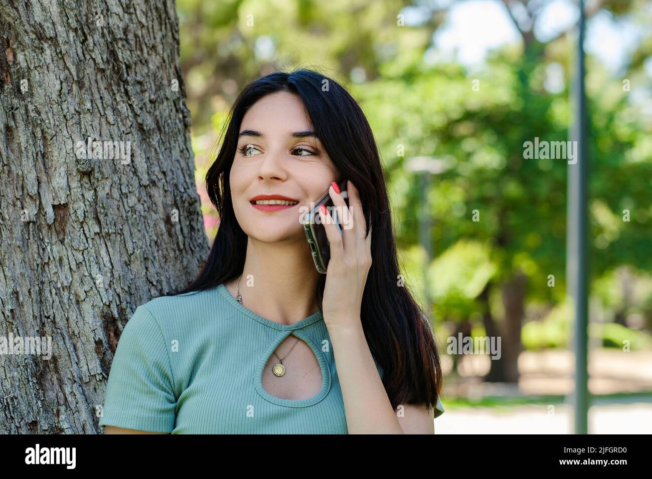 Schöne Brünette Frau in türkisfarbenem T-Shirt im Stadtpark, im Freien im Gespräch auf dem Handy mit Freunden oder Freund mit einem Lächeln. Stockfoto