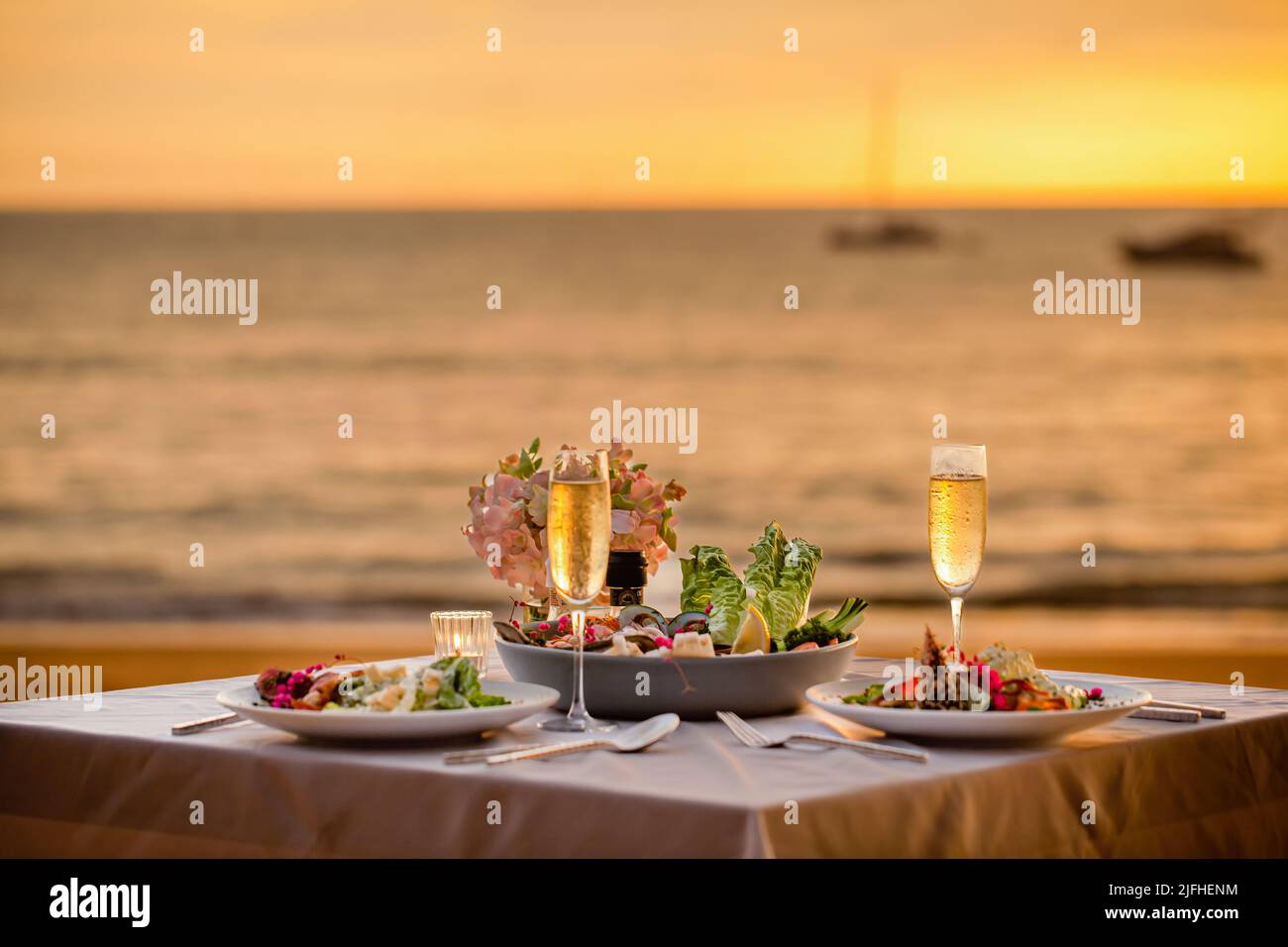 Romantisches Abendessen bei Sonnenuntergang am Strand. Tisch-Flitterwochen-Set für zwei Personen mit luxuriösem Essen, Gläsern Champagner-Getränken im Restaurant mit Meerblick und Yachten Stockfoto