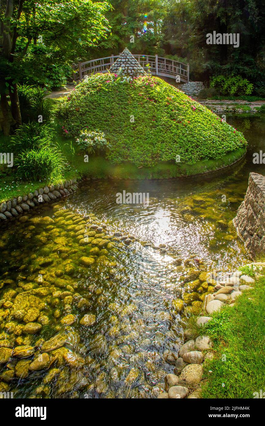 Der Teich mit Steinbänken und einer Art Azaleen Büschen Pyramide und japanische Brücke dahinter und Sonne glänzte in Wasserspiegelungen in Albert Kahn Stockfoto