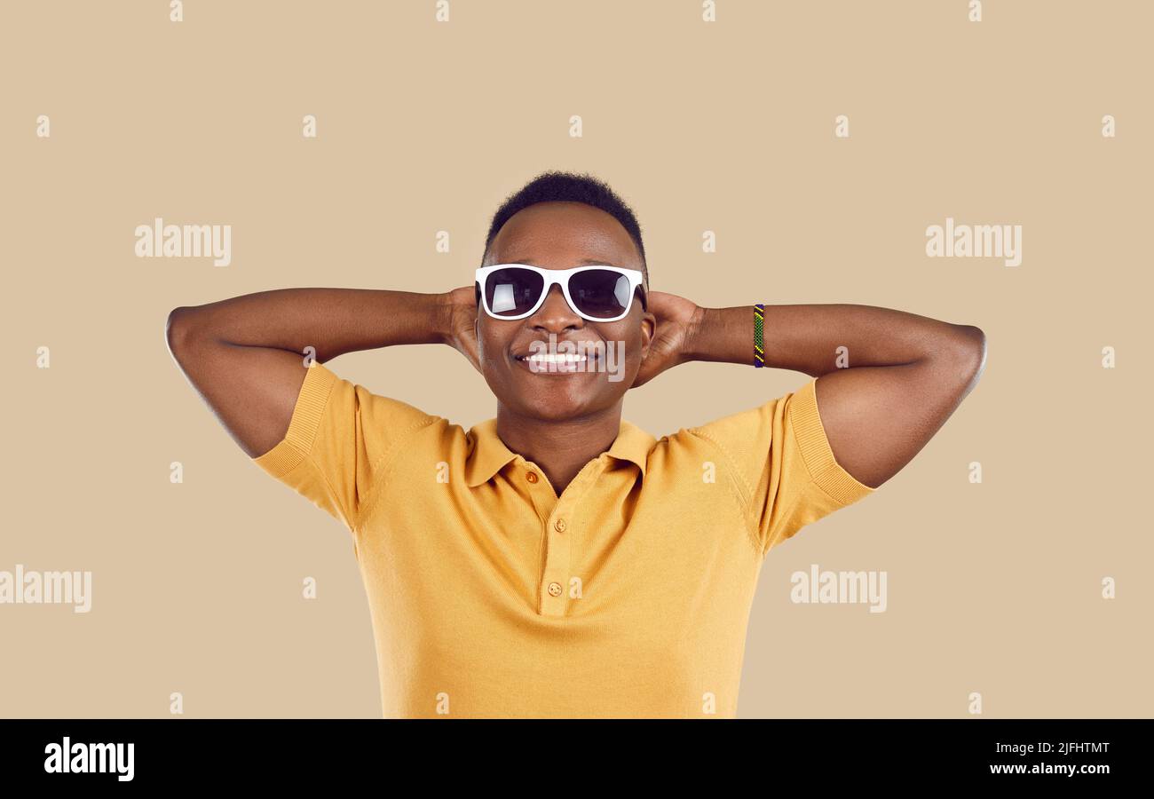 Cooler, glücklicher afroamerikanischer Mann in einer Sonnenbrille, der die Hände hinter dem Kopf auf hellbeigem Hintergrund hält. Stockfoto