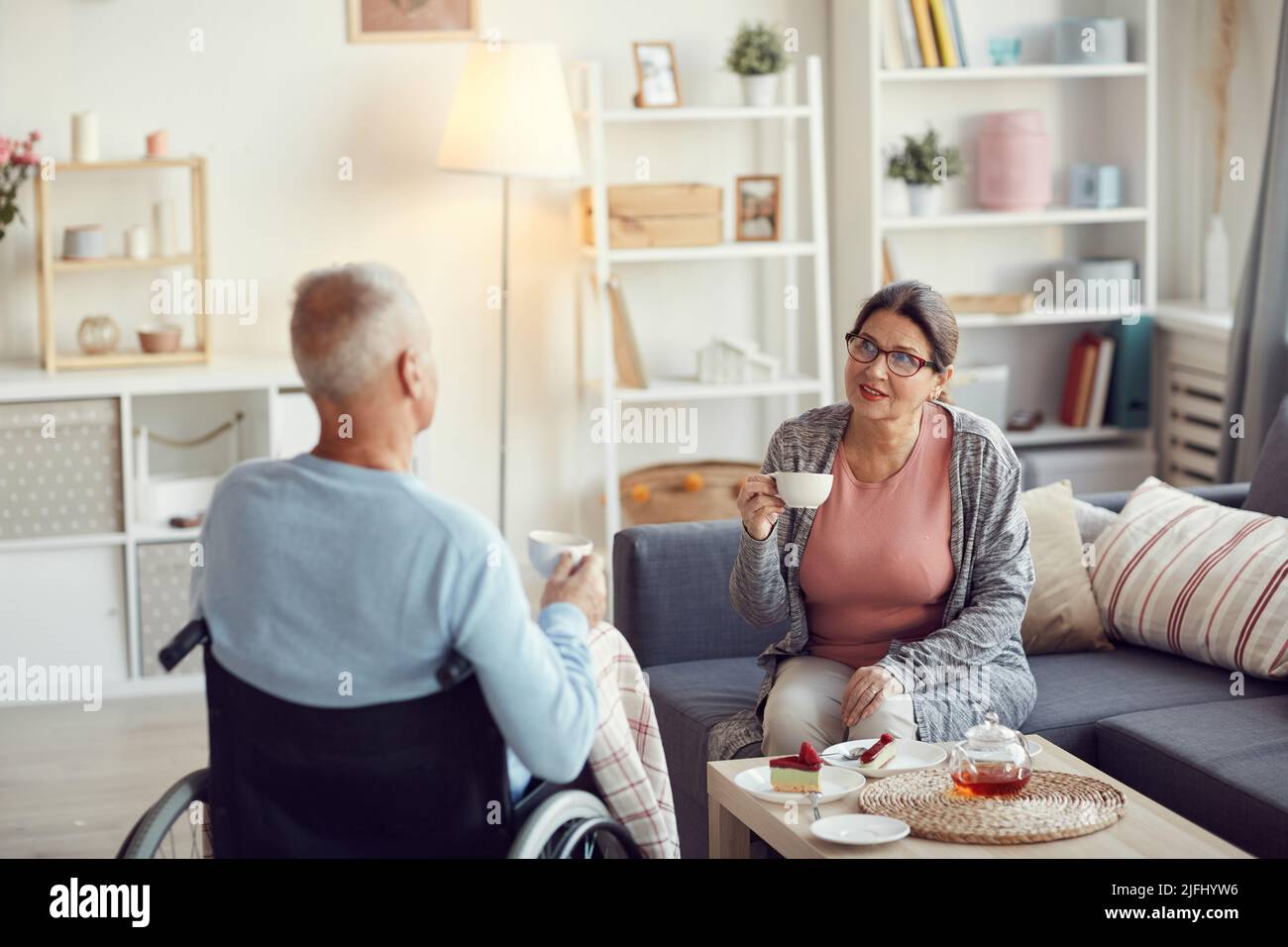 Inhalt ältere Frau im Homewear, die auf dem Sofa sitzt und Tee trinkt, während sie während der Kommunikation einen behinderten Ehemann anlächelt Stockfoto Inhalt ältere Frau im Homewear, die auf dem Sofa sitzt und Tee trinkt, während sie während der Kommunikation einen behinderten Ehemann anlächelt Stockfoto