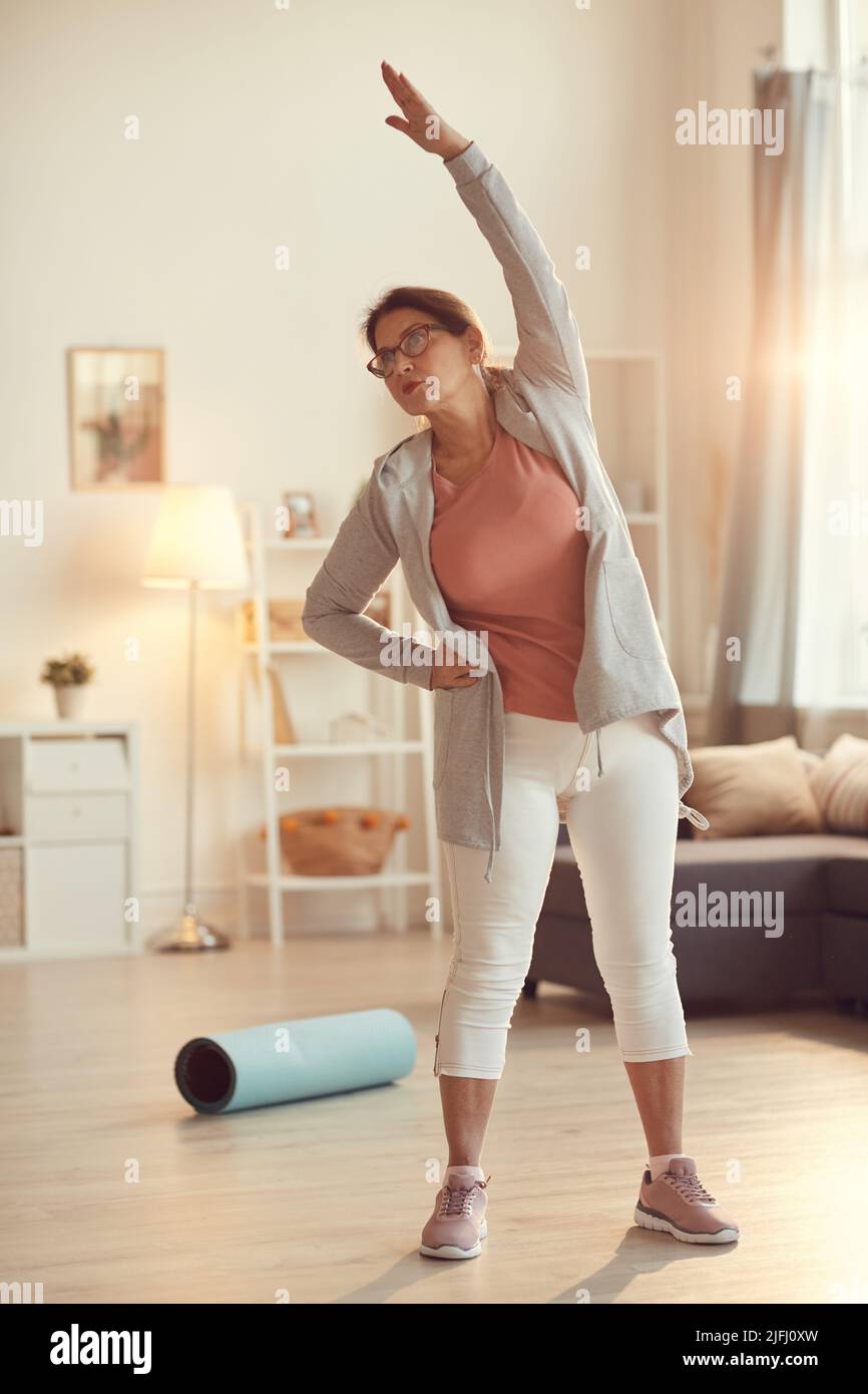 Seriöse, zielgerichtete ältere Frau in Leggings, die im Wohnzimmer steht und alleine Seitenbeugeübungen macht Stockfoto