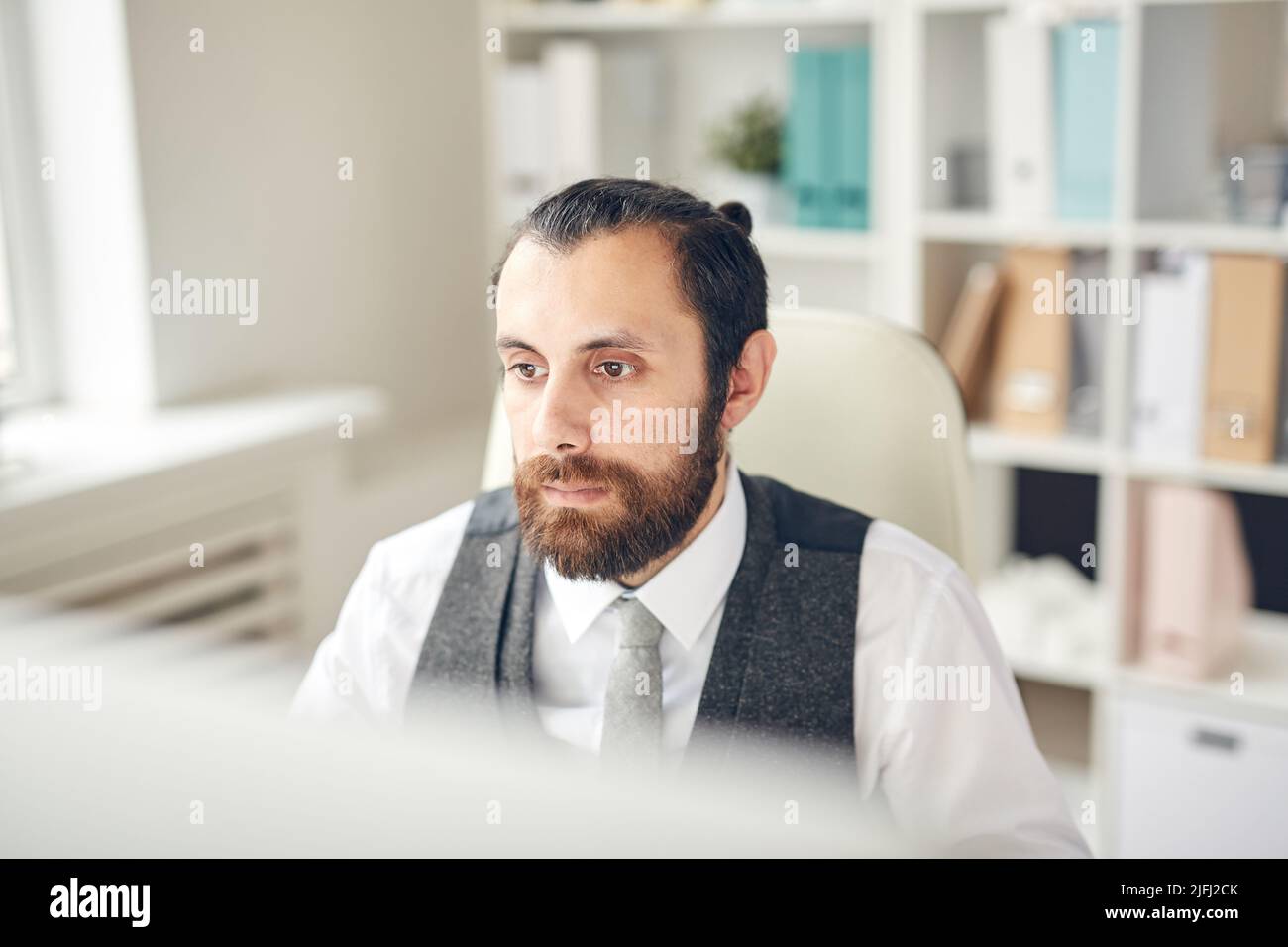 Ernsthafter junger bärtiger Mann in Weste, der vor dem Computermonitor sitzt und die Website des Unternehmens sieht Stockfoto