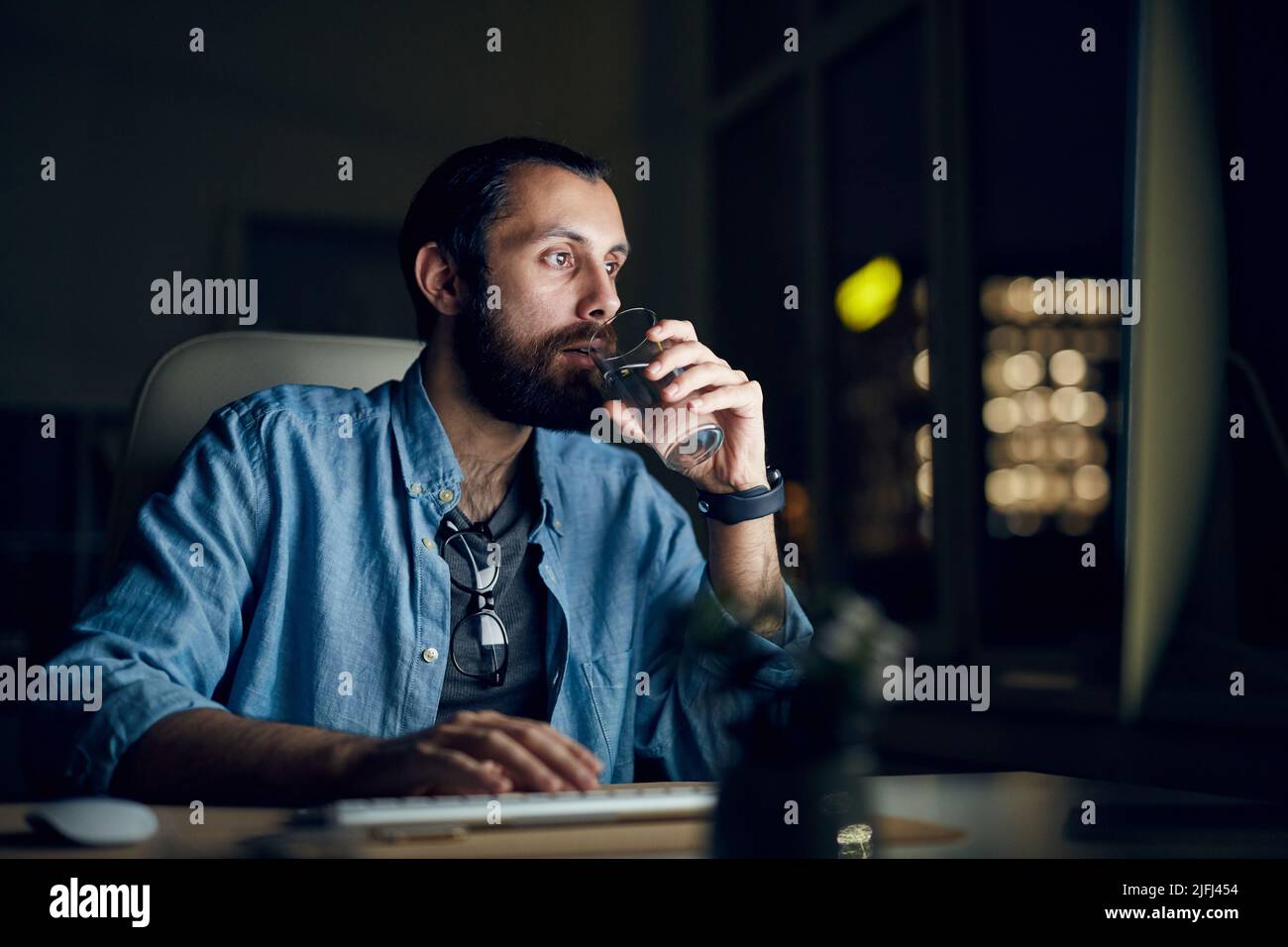 Seriös nachdenklicher junger bärtiger Mann in legerem Hemd, der im Nachtbüro sitzt und Wasser trinkt, während er den Computer benutzt Stockfoto