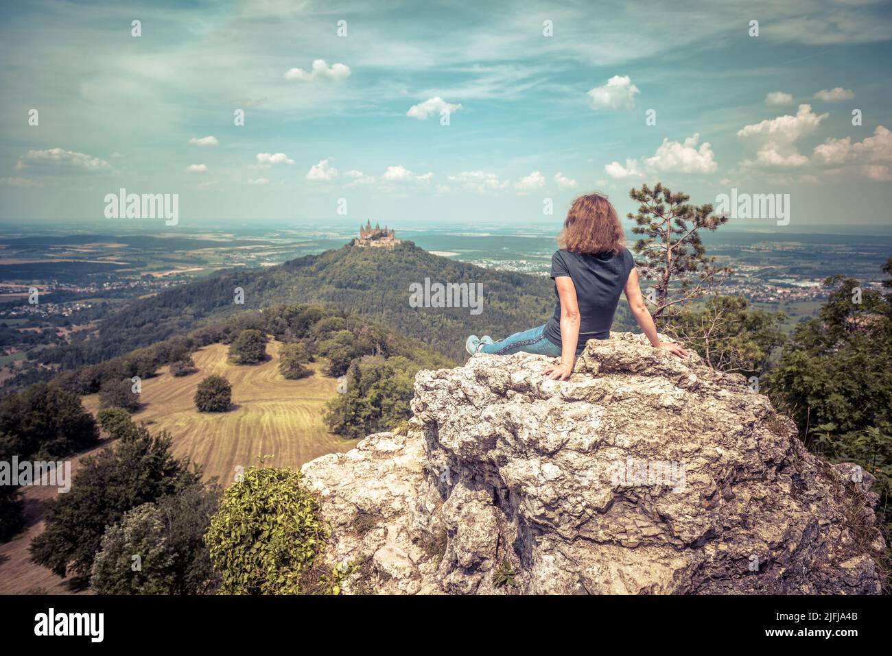 Eine Wandererin, die auf einer Klippe sitzt, blickt auf die Burg Hohenzollern auf dem Berggipfel in Deutschland. Erwachsene Mädchen reist im Sommer in den Schwäbischen Alpen. Konzept von yo Stockfoto