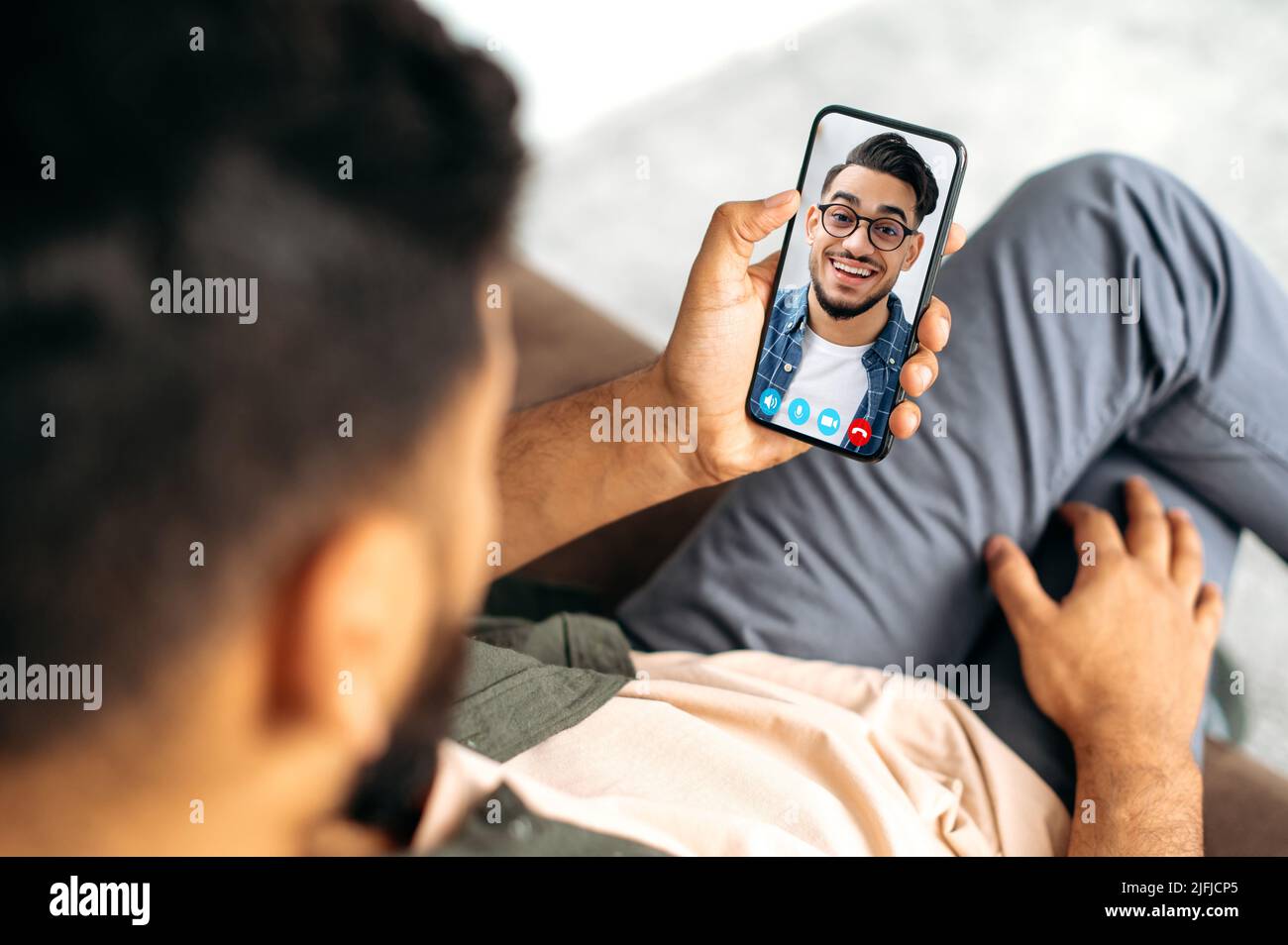 Blick auf ein Telefon mit indischen oder arabischen Kerl. Zwei Männer sprechen auf einer Online-Videokonferenz mit einer Anwendung und einem Smartphone, sind in einem Abstand voneinander, lächelnd freundlich Stockfoto