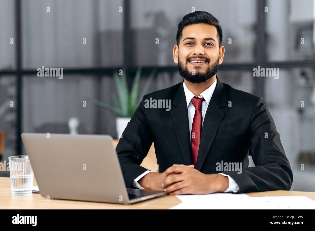 Porträt eines positiven jungen männlichen selbstbewussten arabischen oder indischen Geschäftsmannes, erfolgreicher Unternehmer in einem Anzug, sitzen an einem Schreibtisch mit Laptop in einem modernen kreativen Büro, blickt auf die Kamera, lächeln freundlich Stockfoto