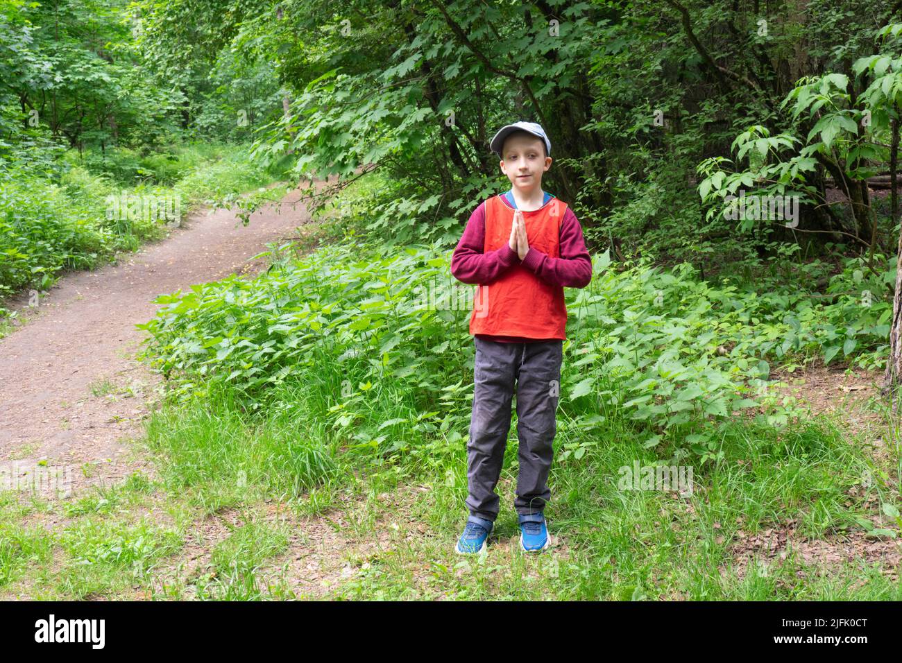 Der Junge in einem roten Hemd, einer grauen Hose und einer Denim-Mütze hält seine Hände in einer namastigen Grußgeste vor sich Stockfoto