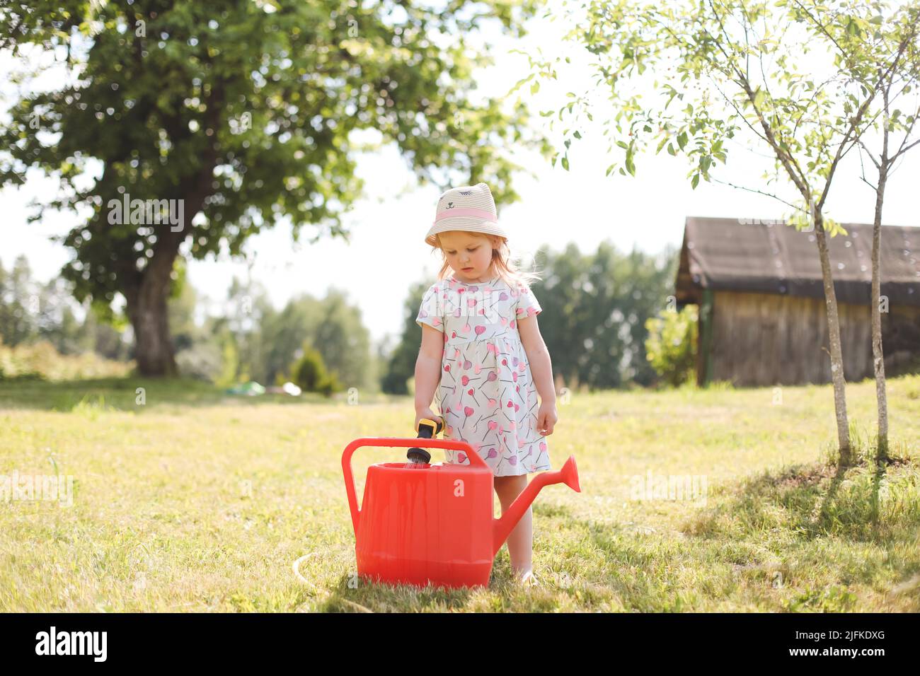 Ein kleines niedliches Baby Mädchen 3-4 Jahre alt in einem Kleid, das die Pflanzen aus einer Gießkannen im Garten wässert. Kinder haben Spaß beim Gartenarbeiten auf einem hellen sonnigen Stockfoto