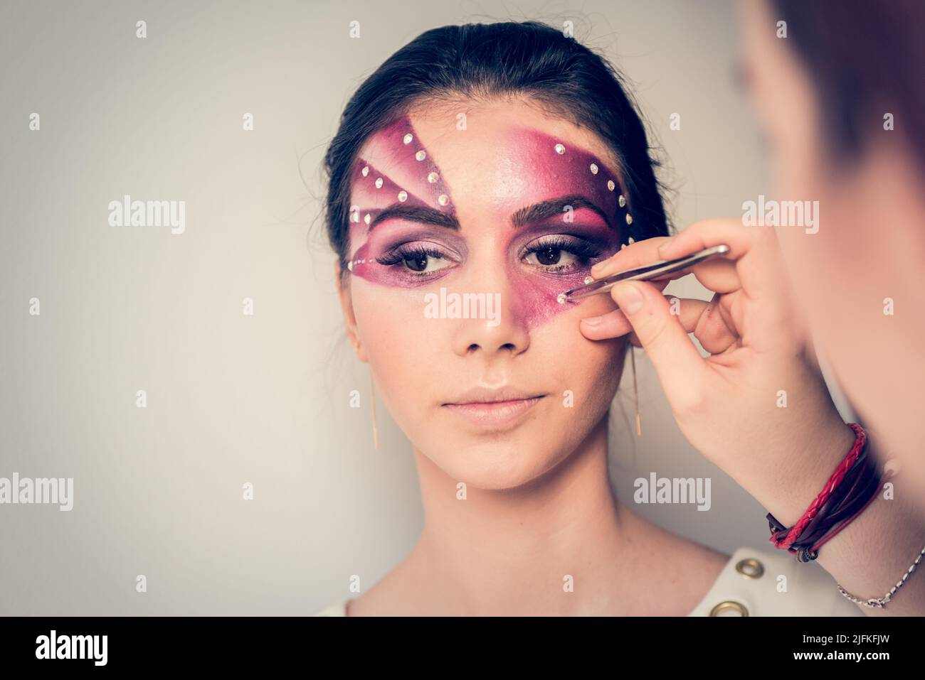 Nachdem die Make-up-Künstlerin ihr Gesicht mit Purpur bemalt hat, applyiert sie Kristalle auf das Gesicht eines jungen Familienmodells mit einer Pinzette Stockfoto