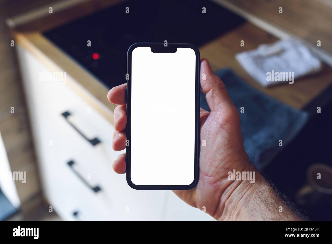 Home Automation und Internet of Things Konzept, männliche Hand hält Smartphone mit leerem Mockup-Bildschirm vor dem Küchentischtisch, selektiver Fokus Stockfoto