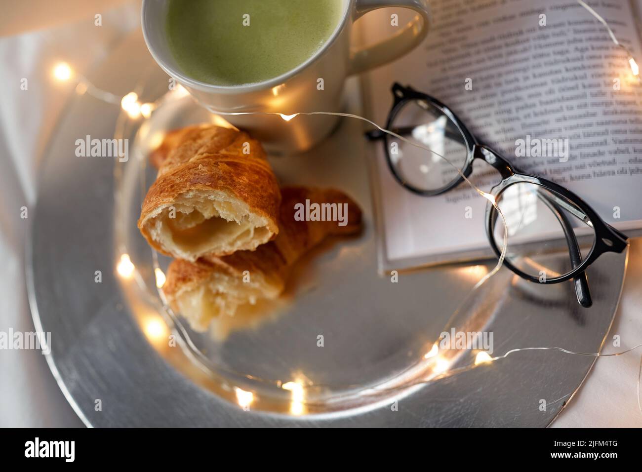 Croissants, Matcha-Tee, Buch und Gläser im Bett Stockfoto