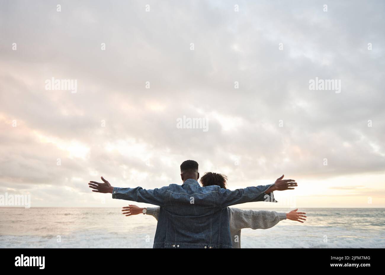 Junges multiethnisches Paar, das mit erhobenen Armen am Strand steht Stockfoto