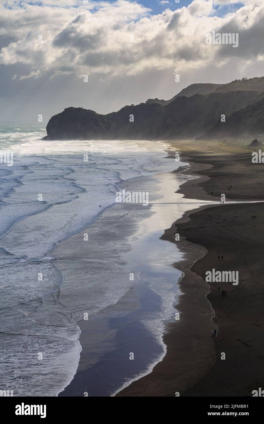Piha, ein schwarzer Sandstrand in Neuseeland, an einem windigen Herbsttag. Sonnenlicht bricht durch die Wolken, um den Te Waha Point zu erleuchten Stockfoto