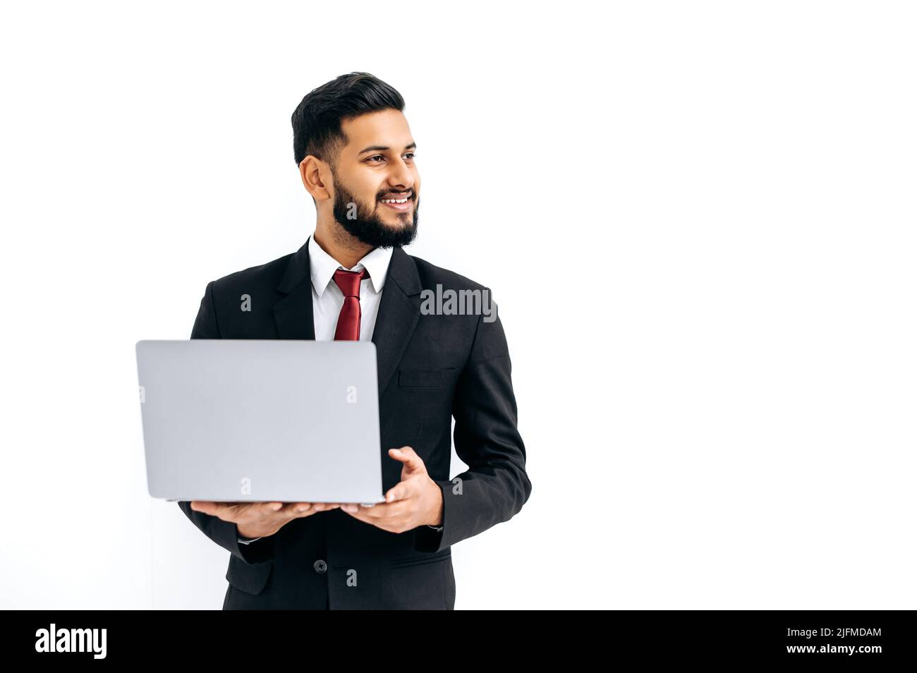 Positiv schöner eleganter indischer oder arabischer junger Mann im Business-Anzug, männlicher Manager, mit offenem Laptop in den Händen, zur Seite schauend, auf isoliertem weißen Hintergrund stehend, lächelnd freundlich. Speicherplatz kopieren Stockfoto