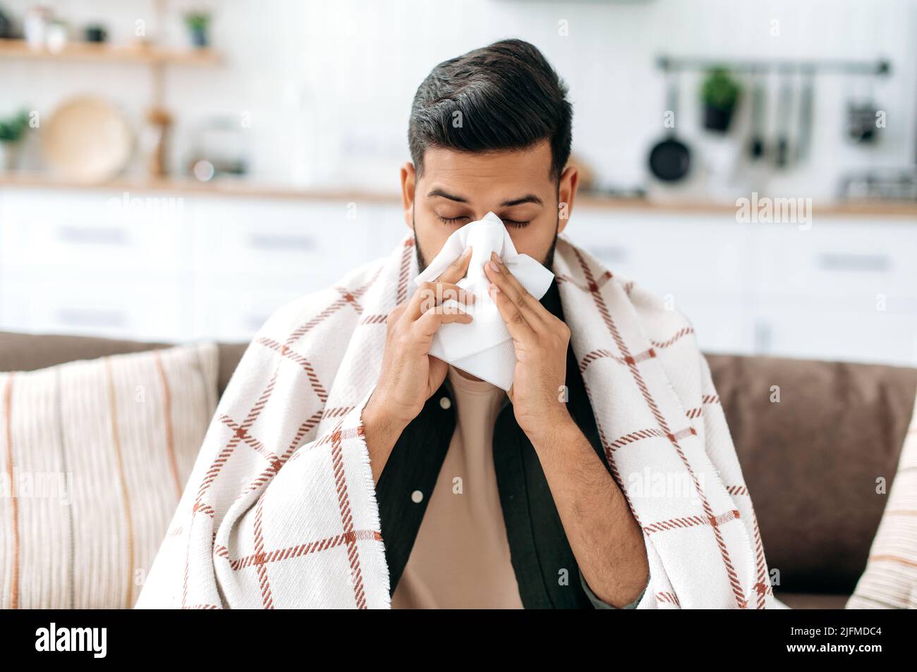 Ungesunder inder oder araber sitzt zu Hause auf dem Sofa im Wohnzimmer unter Plaid, hält Serviette in der Nähe der Nase, niest, leidet an laufender Nase, fühlt sich unwohl, braucht eine Behandlung und einen Arztbesuch Stockfoto