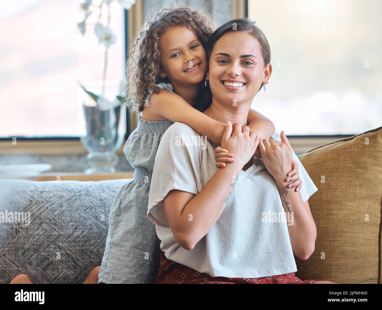 Porträt eines kleinen Mädchens, das ihre Mutter von hinten umarmt und Liebe und Zuneigung zeigt, während sie zu Hause auf der Couch sitzt. Süßer Moment zwischen der Mutter Stockfoto