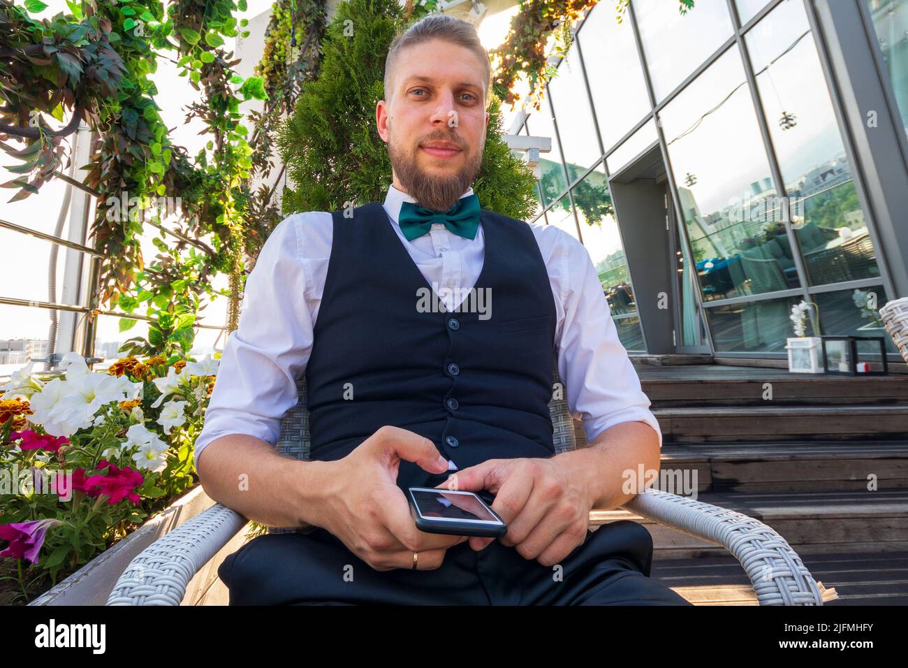 Ein junger, selbstbewusster Mann in einem weißen Hemd, einer schwarzen Weste mit einem Smartphone in den Händen schaut auf die Kamera, während er im Penthouse sitzt Stockfoto