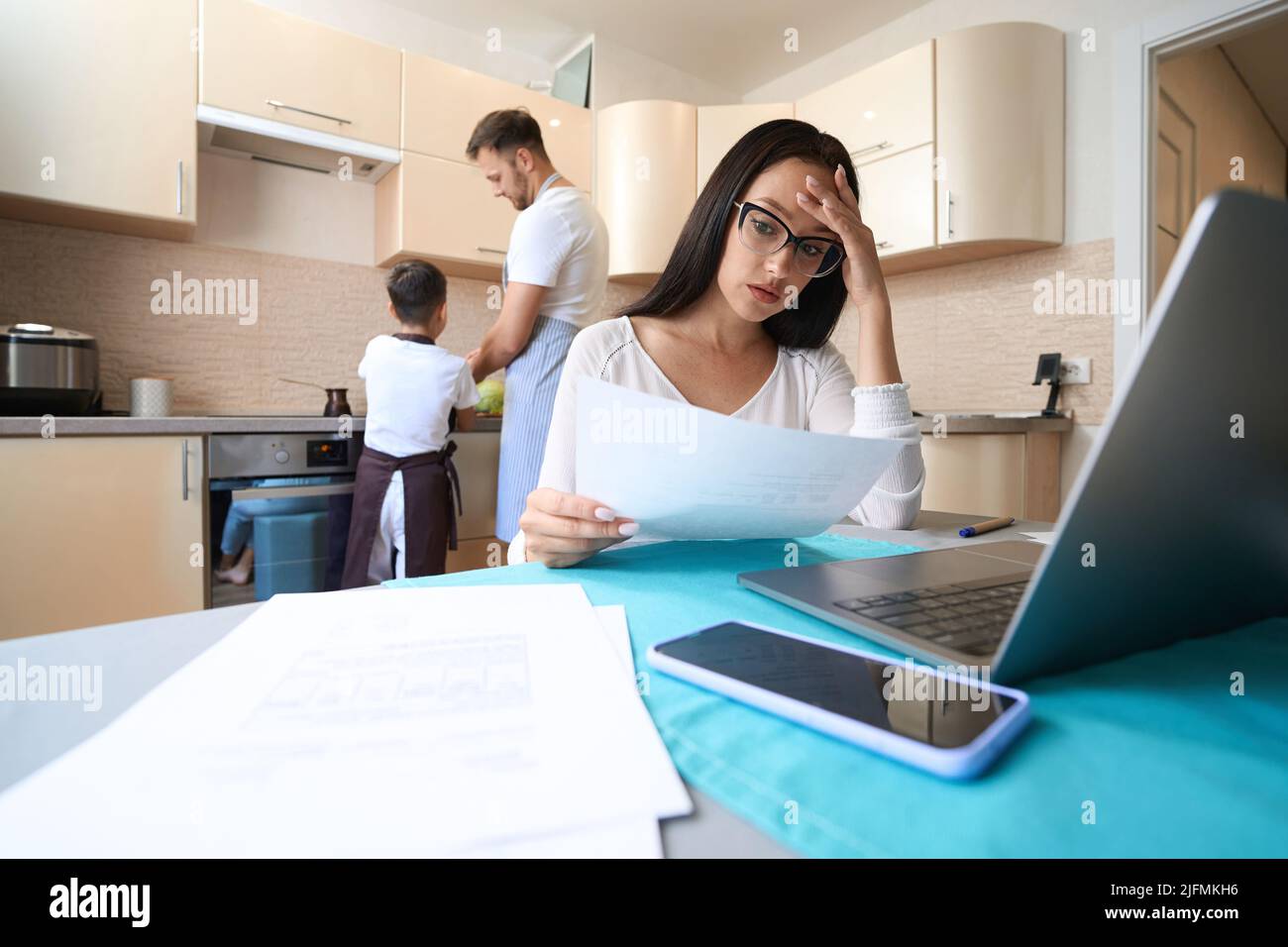 Müde Frau, die Papier nachprüfte, während sie in der Küche an einem Laptop arbeitete Stockfoto