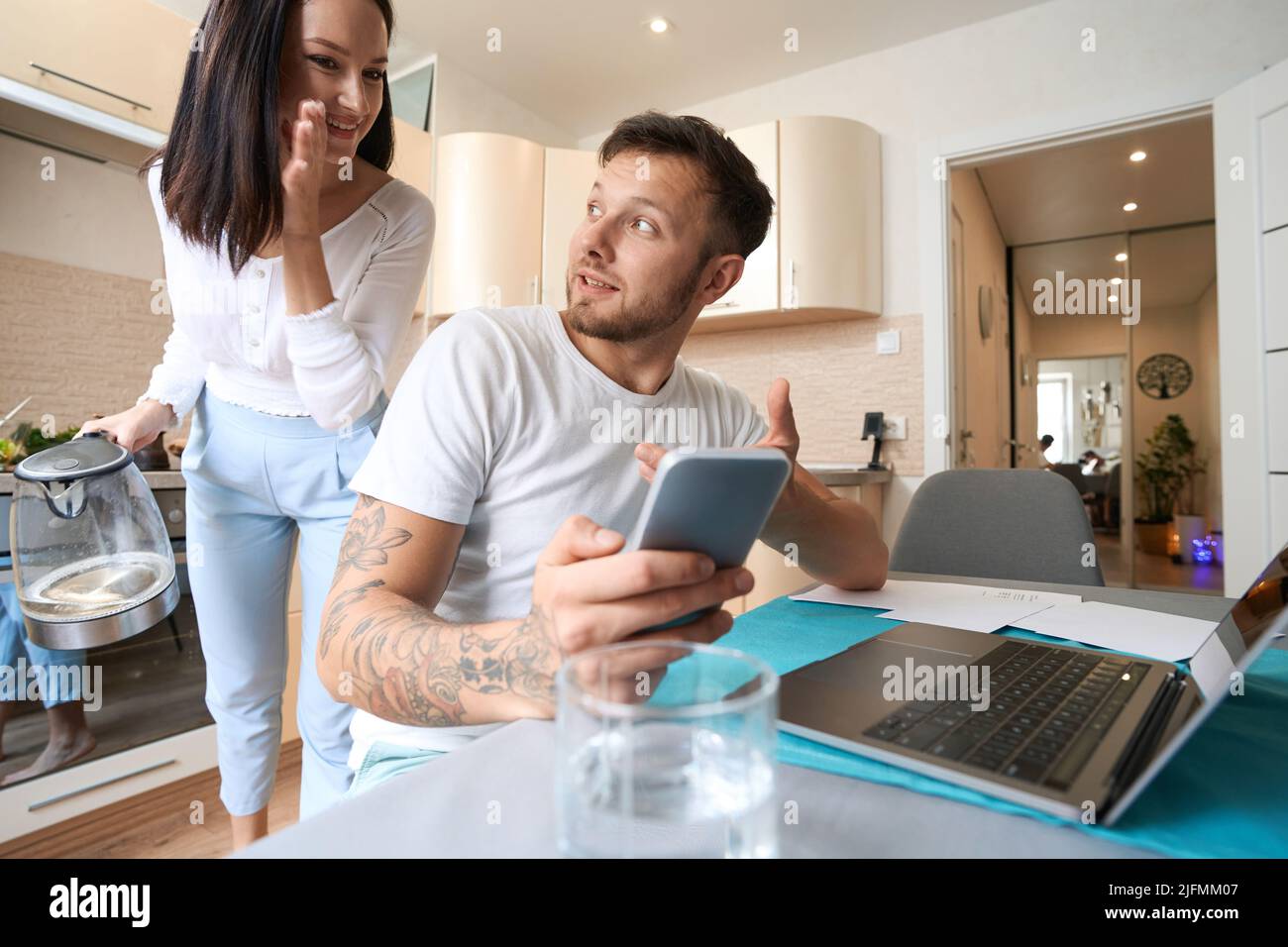 Frau gibt dem Ehemann Ratschläge, arbeitet mit dem Smartphone am Laptop Stockfoto
