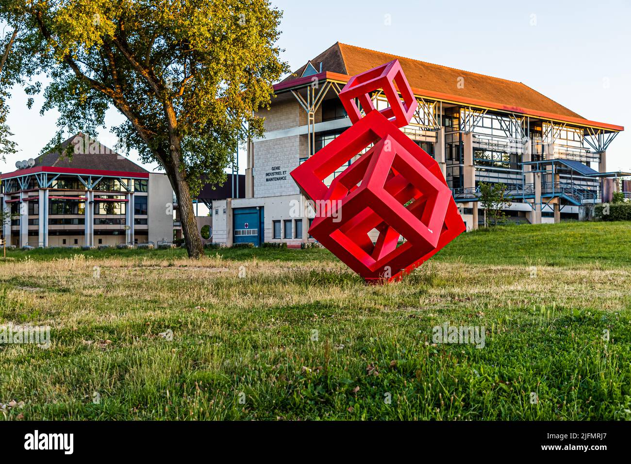 Eine rote Skulptur aus ineinandergreifenden Würfeln steht auf dem Rasen vor dem IUT-Institut der Universität Burgund in Chalon-sur-Saône, Frankreich. Ein Schild am Gebäude markiert die „Zone Industrielle et Maintenance“. Stockfoto