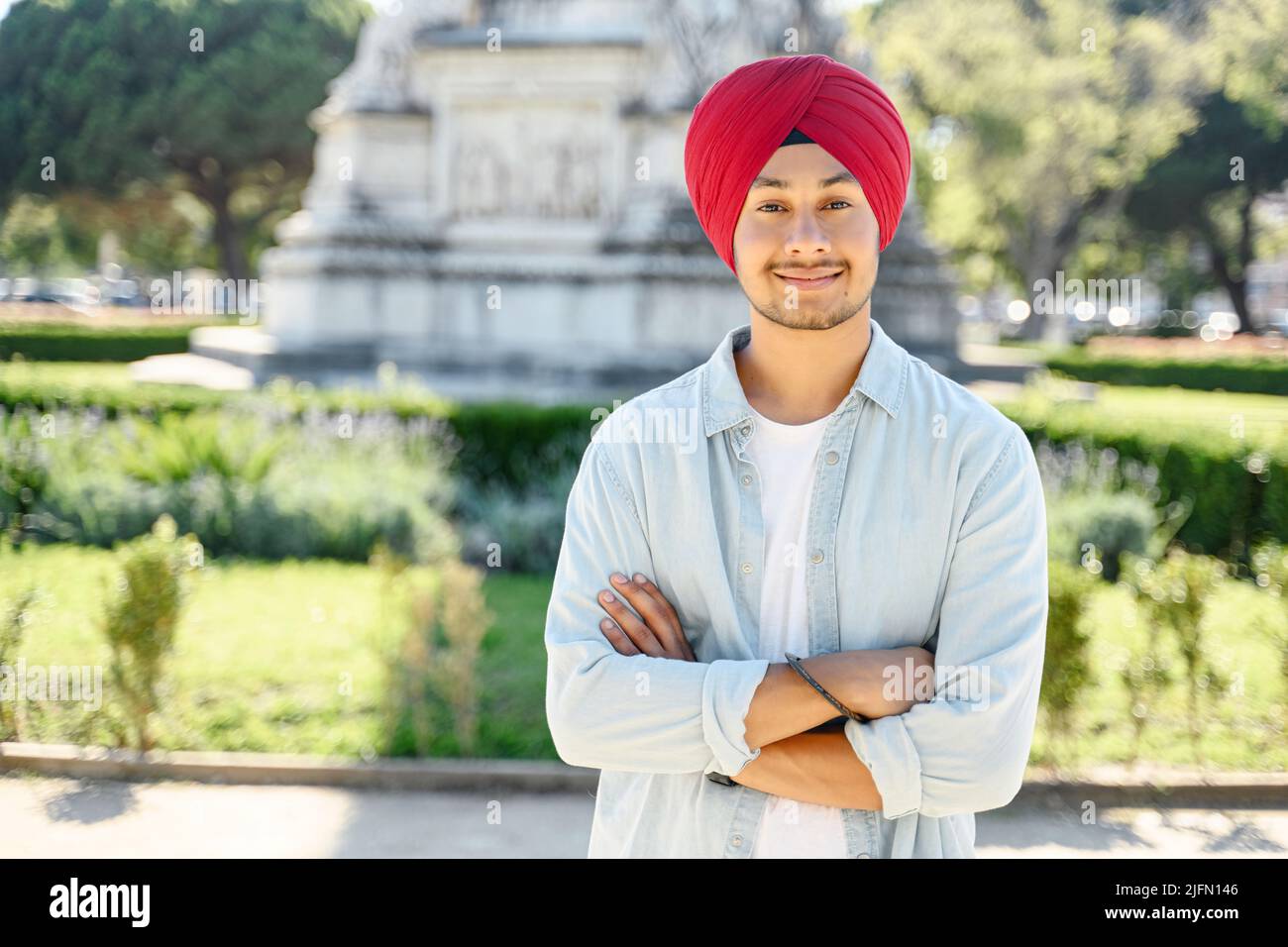 Ein hübscher, lächelnder junger Inder mit traditionellem roten Turban steht draußen mit gefalteten Armen. Stolzer, selbstbewusster sikh-Mann mit gekreuzten Armen und Blick auf die Kamera, die im Stadtbild steht Stockfoto
