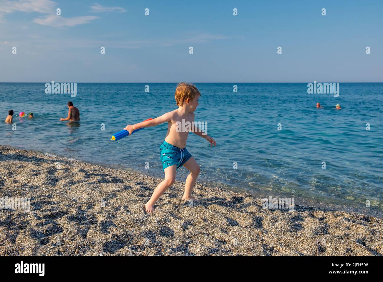 Netter kleiner Junge mit blonden Haaren, der mit seinem Wasserpistole im Meer spielt Stockfoto