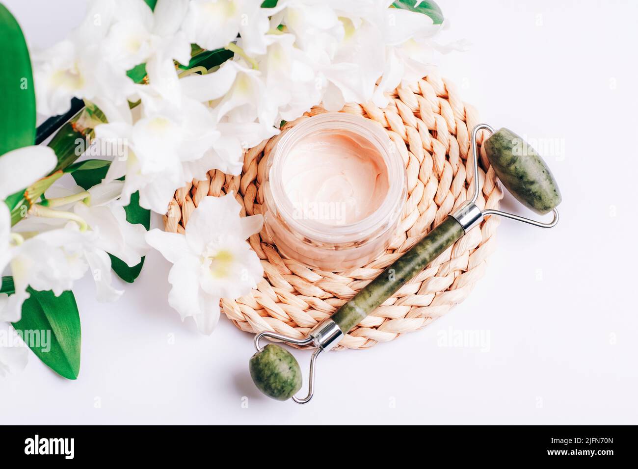 Cremedose und Jade Gesichtswalze mit großen weißen Blüten. Beauty-, Spa- und Wellness-Konzept. Draufsicht, flach liegend Stockfoto